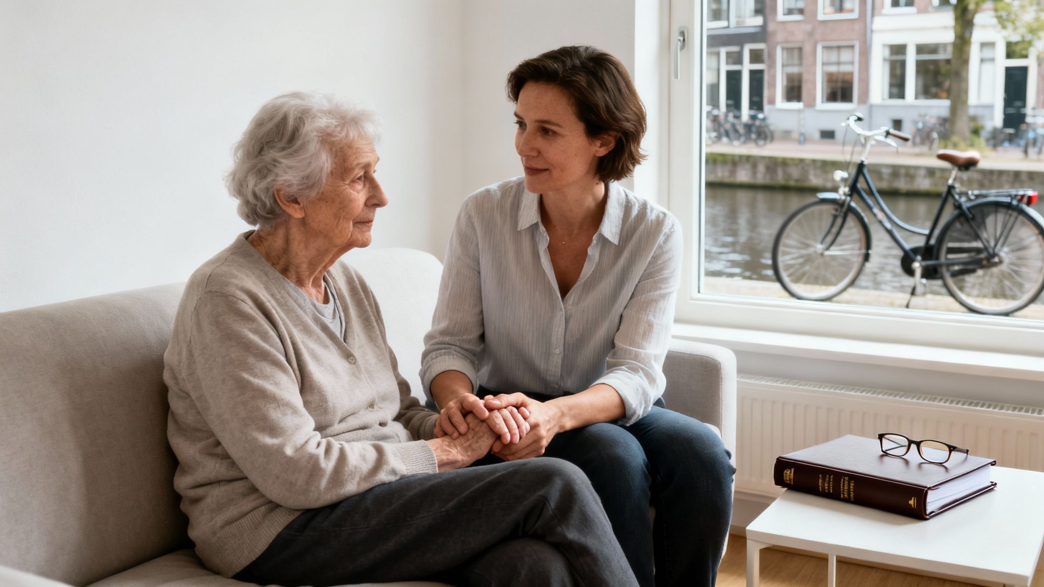 A younger woman holds the hand of an elderly woman, sharing a supportive moment on a sofa.