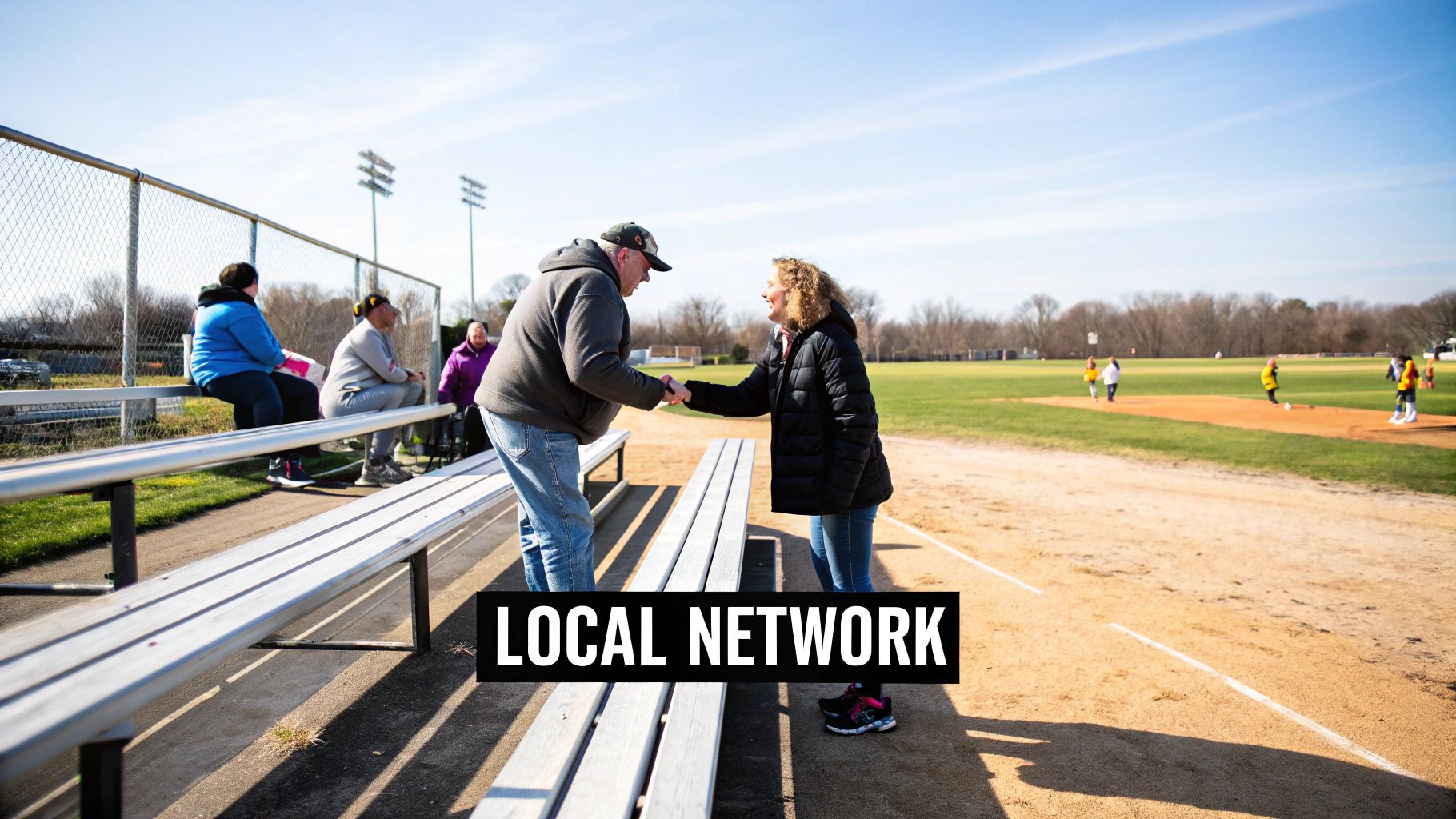 Two adults shake hands on a sunny baseball field near bleachers with other spectators.