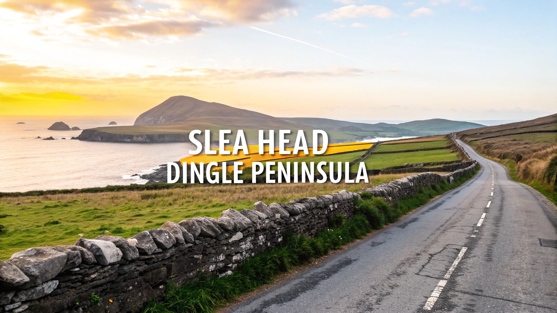 A scenic coastal road with stone walls and green fields leading to the ocean and mountains at sunset.