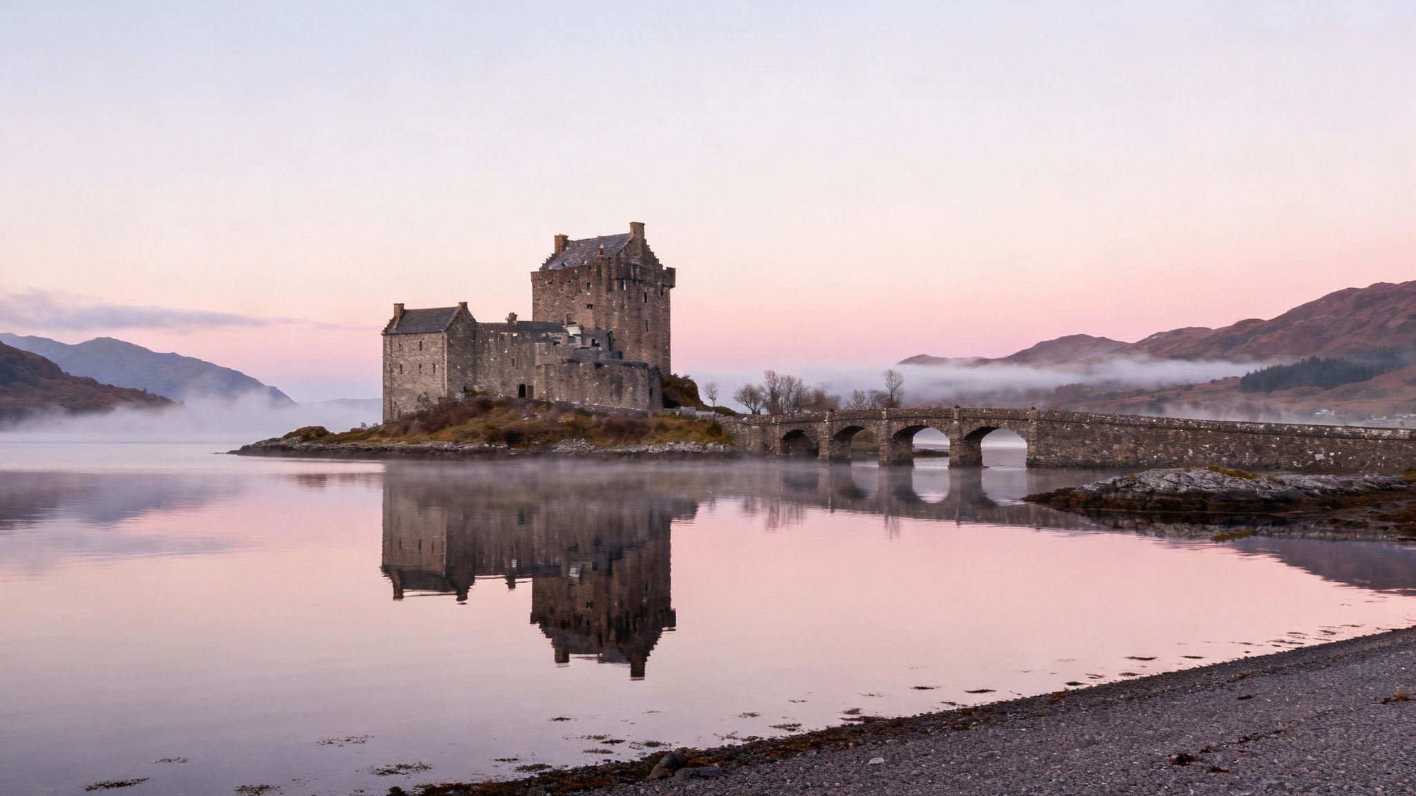 Historic Scottish castle with stone bridge reflected in calm loch waters at misty dawn