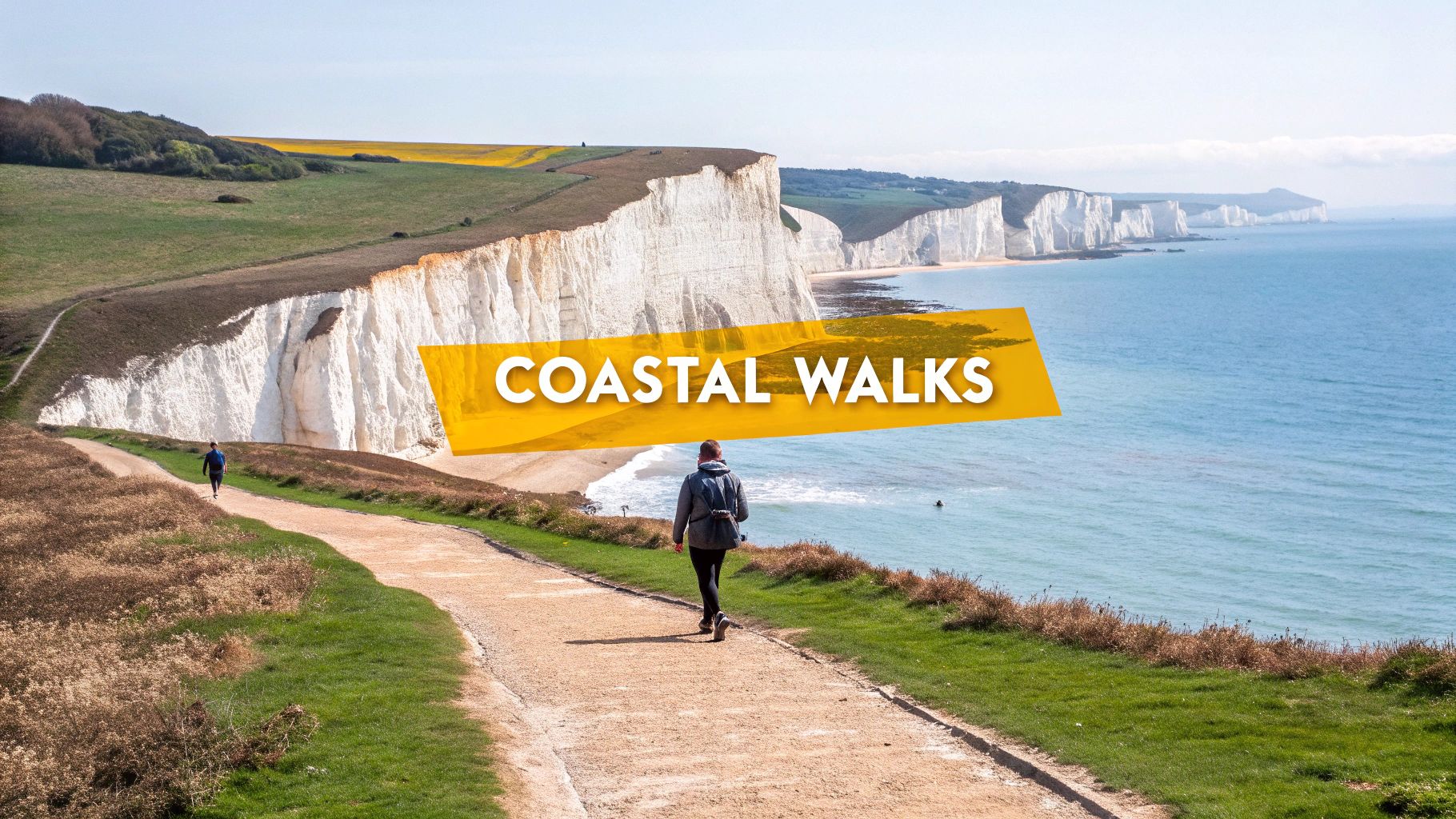 A person walks on a coastal path overlooking white cliffs and the turquoise sea on a sunny day.