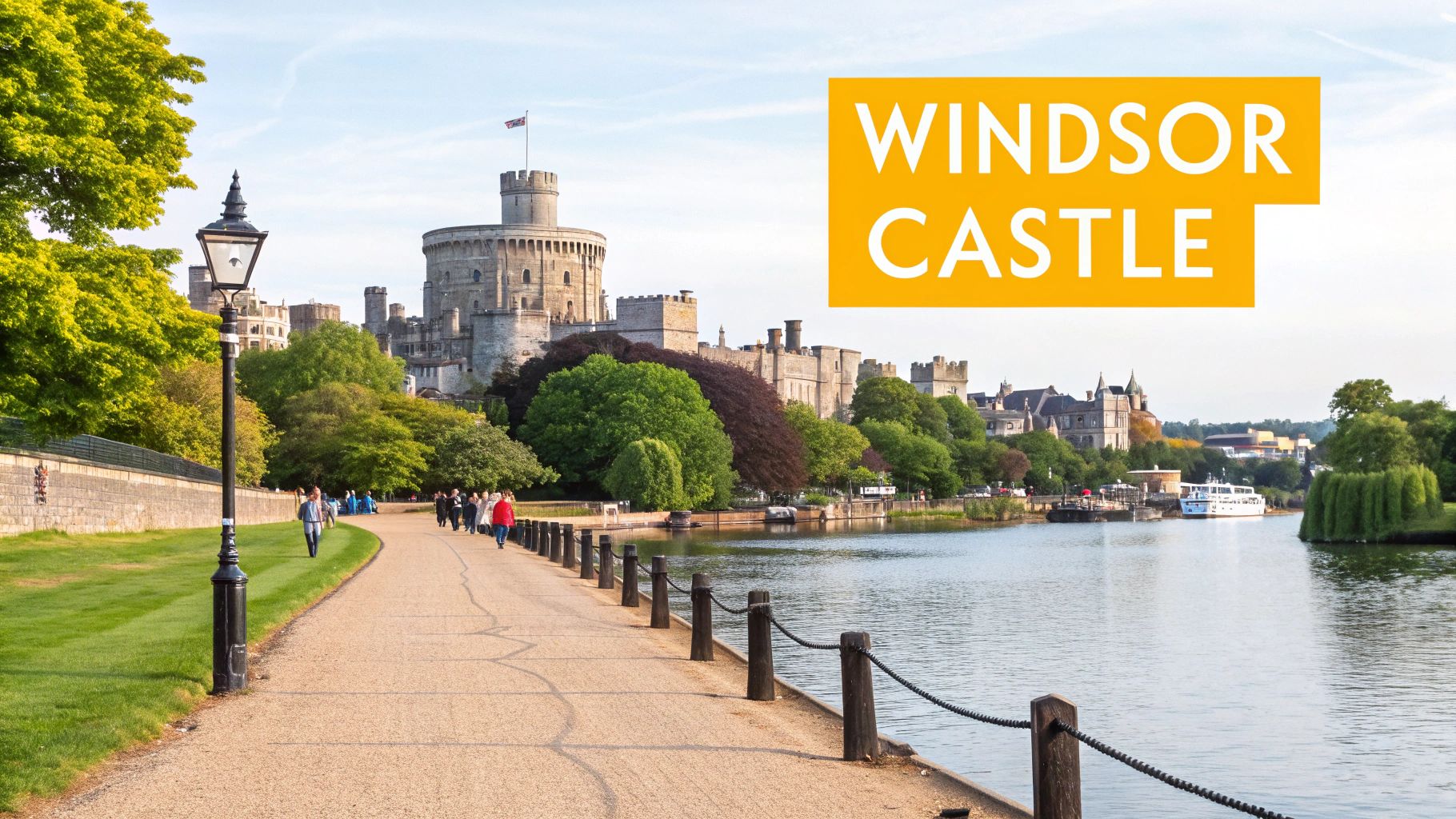 A scenic view of Windsor Castle from across the River Thames, with a riverside path, people, and a lamppost.