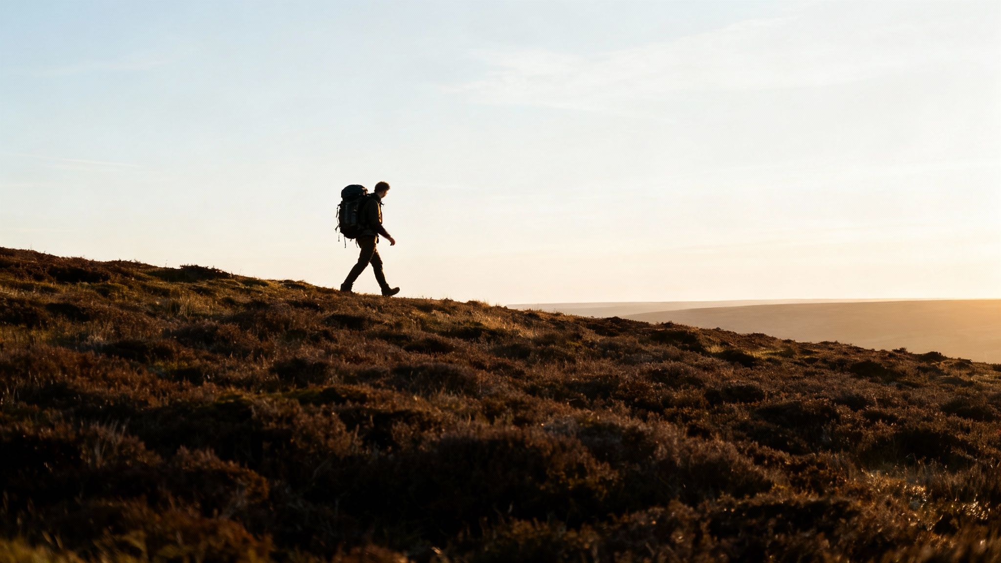 Solo hiker with backpack walking across hillside terrain during golden hour sunset
