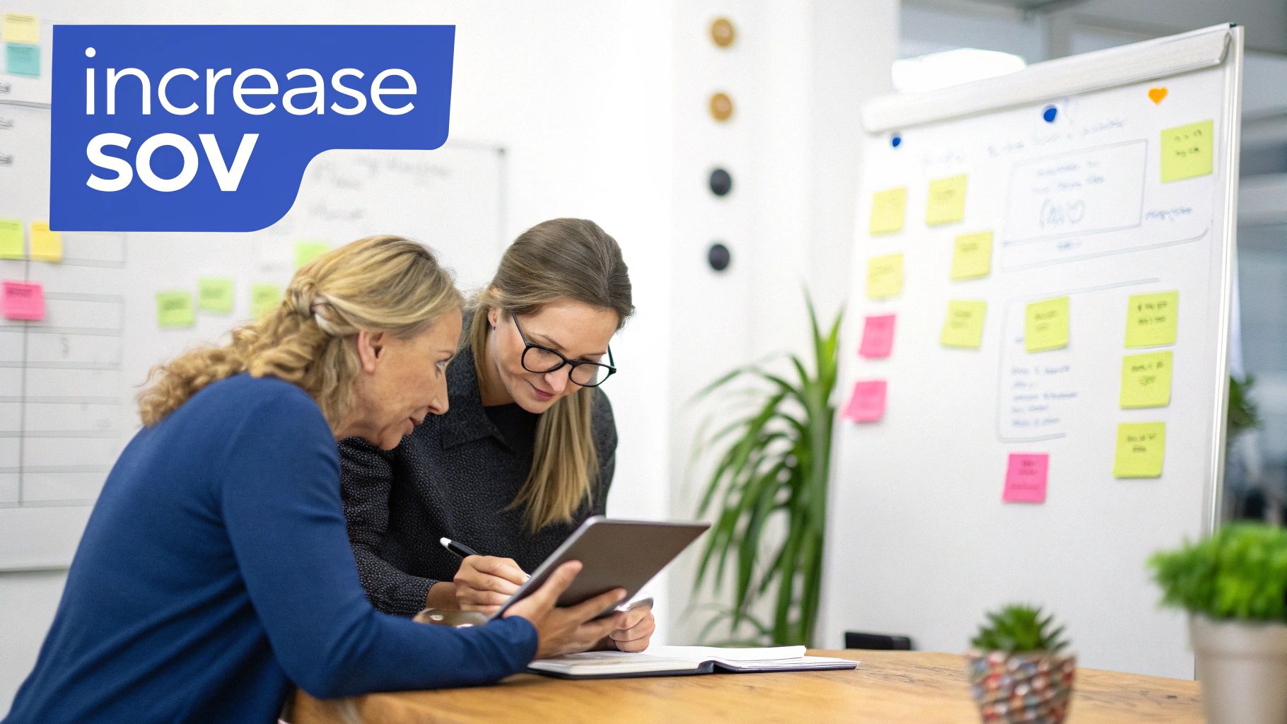 Two businesswomen collaborate on a project, looking at a tablet and taking notes, with a whiteboard in the background.