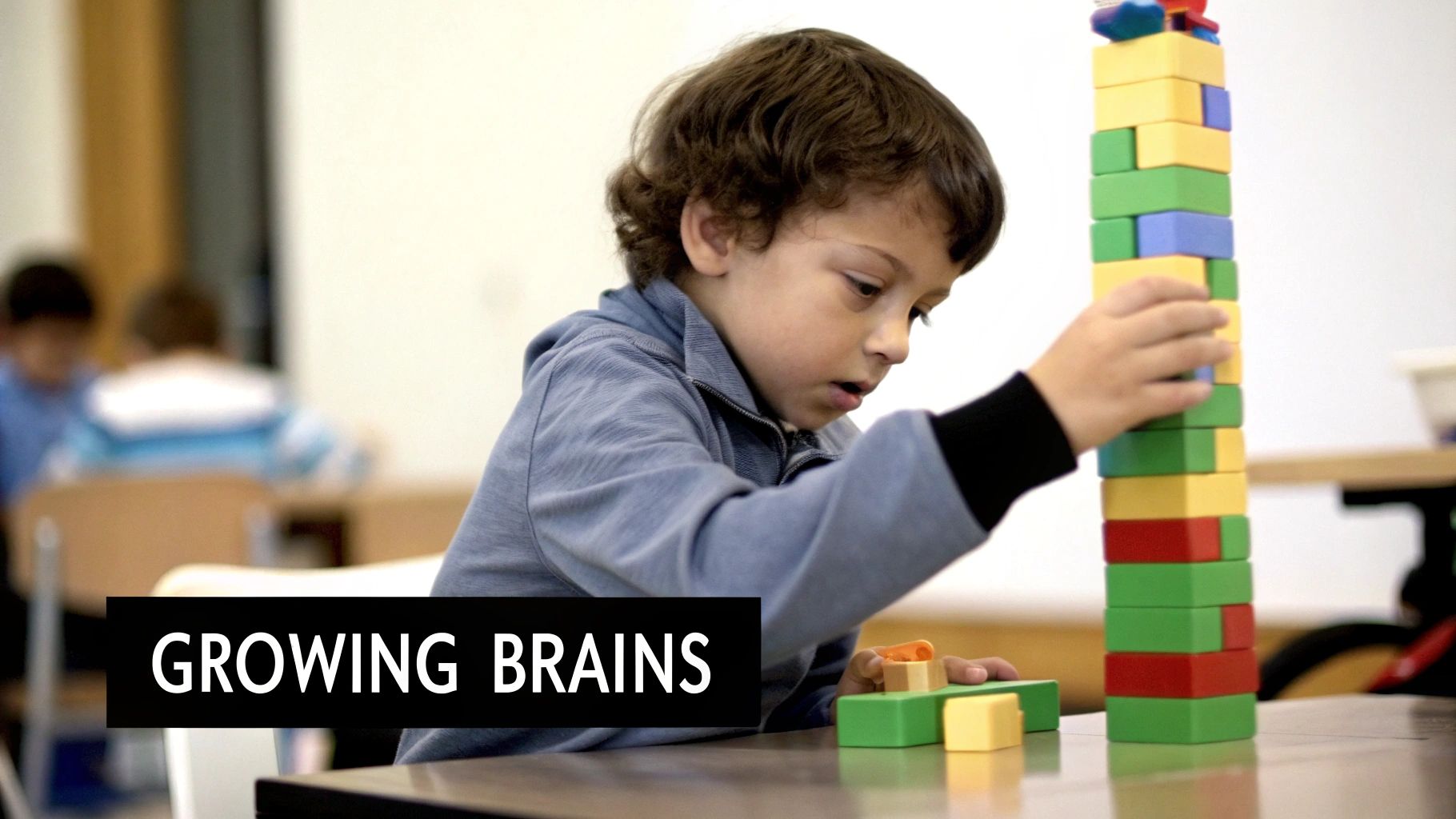 A young boy with curly hair focuses intently while building a tall tower of colorful blocks in a classroom setting.