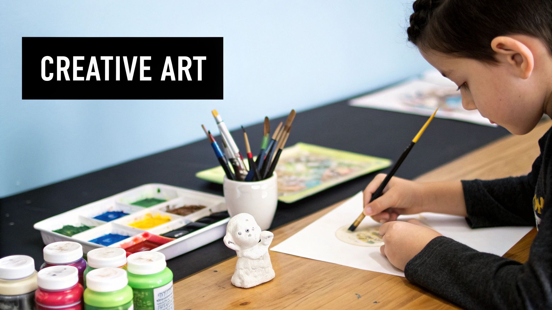 A young child concentrates while painting on paper at a wooden table with various art supplies.