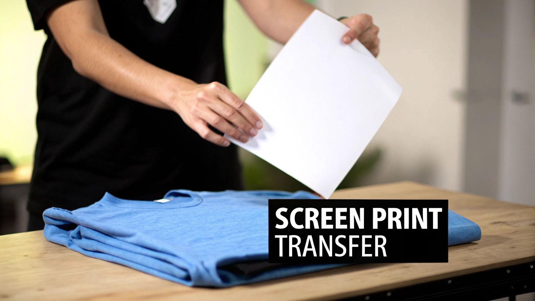 A person holds a blank transfer sheet over a folded blue t-shirt on a wooden table for screen printing.