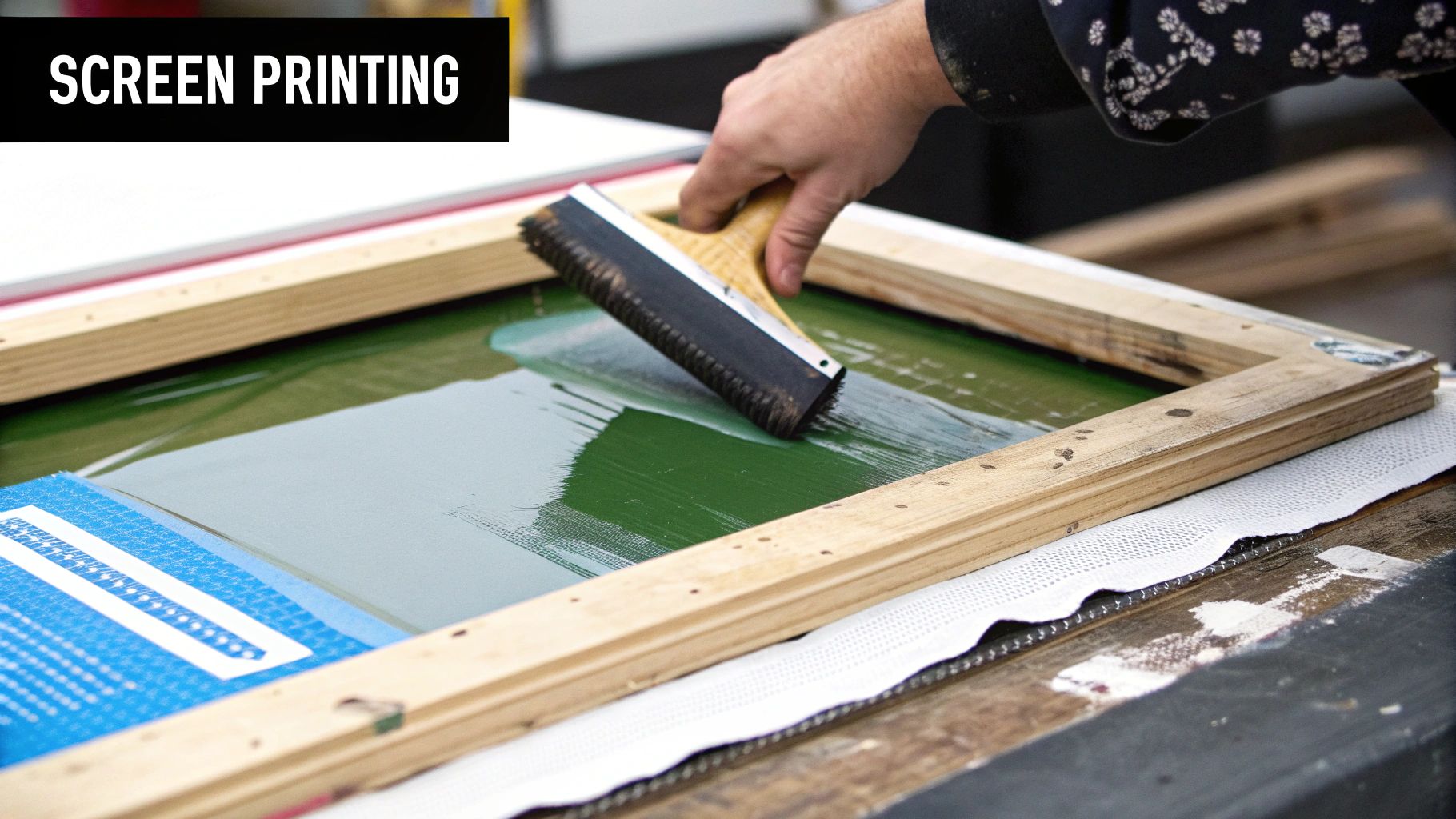 A screen printing press in action, showing a squeegee pushing ink through a mesh screen onto a t-shirt.