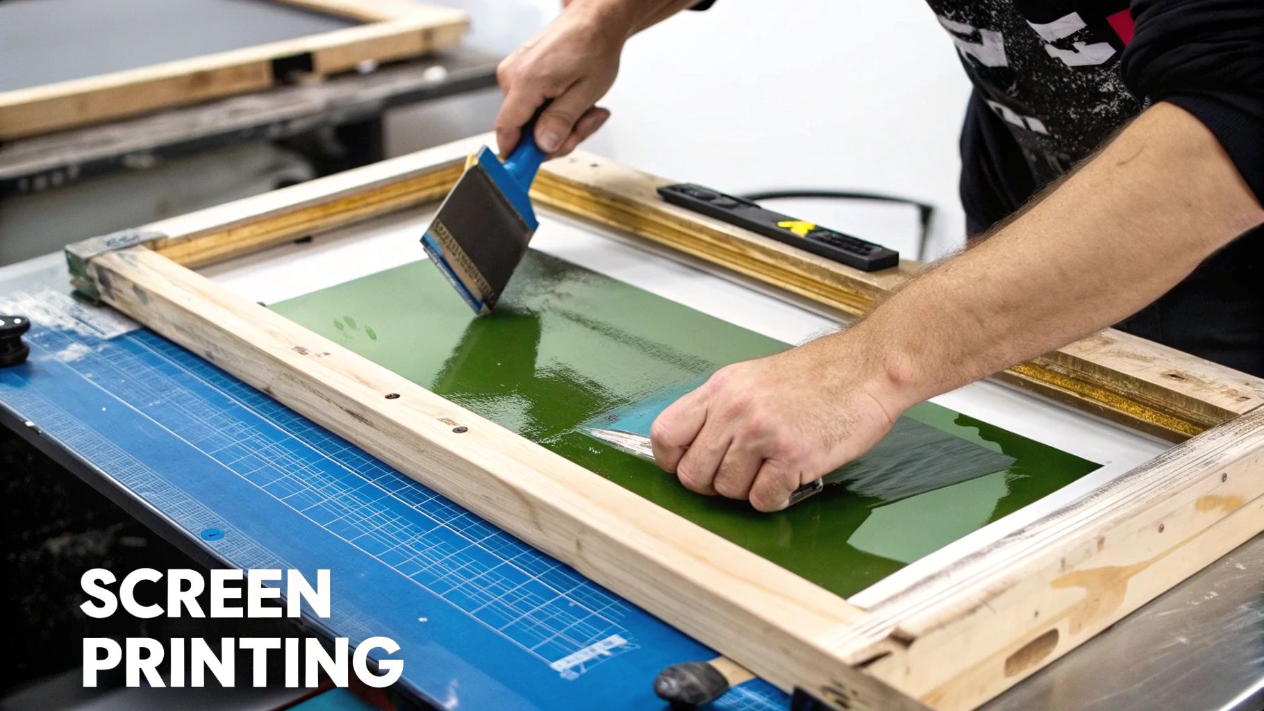 A close-up of a screen printing squeegee pressing vibrant yellow ink through a mesh screen onto a black t-shirt.
