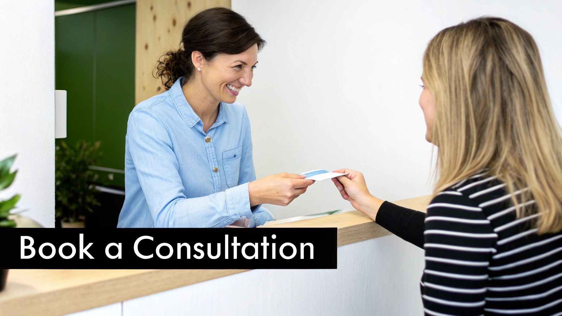 A smiling receptionist hands a card to a client at a counter, suggesting a consultation booking.