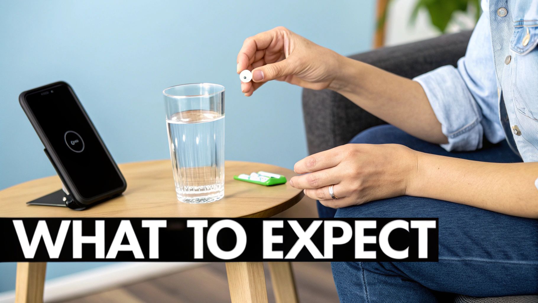 A person holds a small white pill above a glass of water, next to a charging smartphone.
