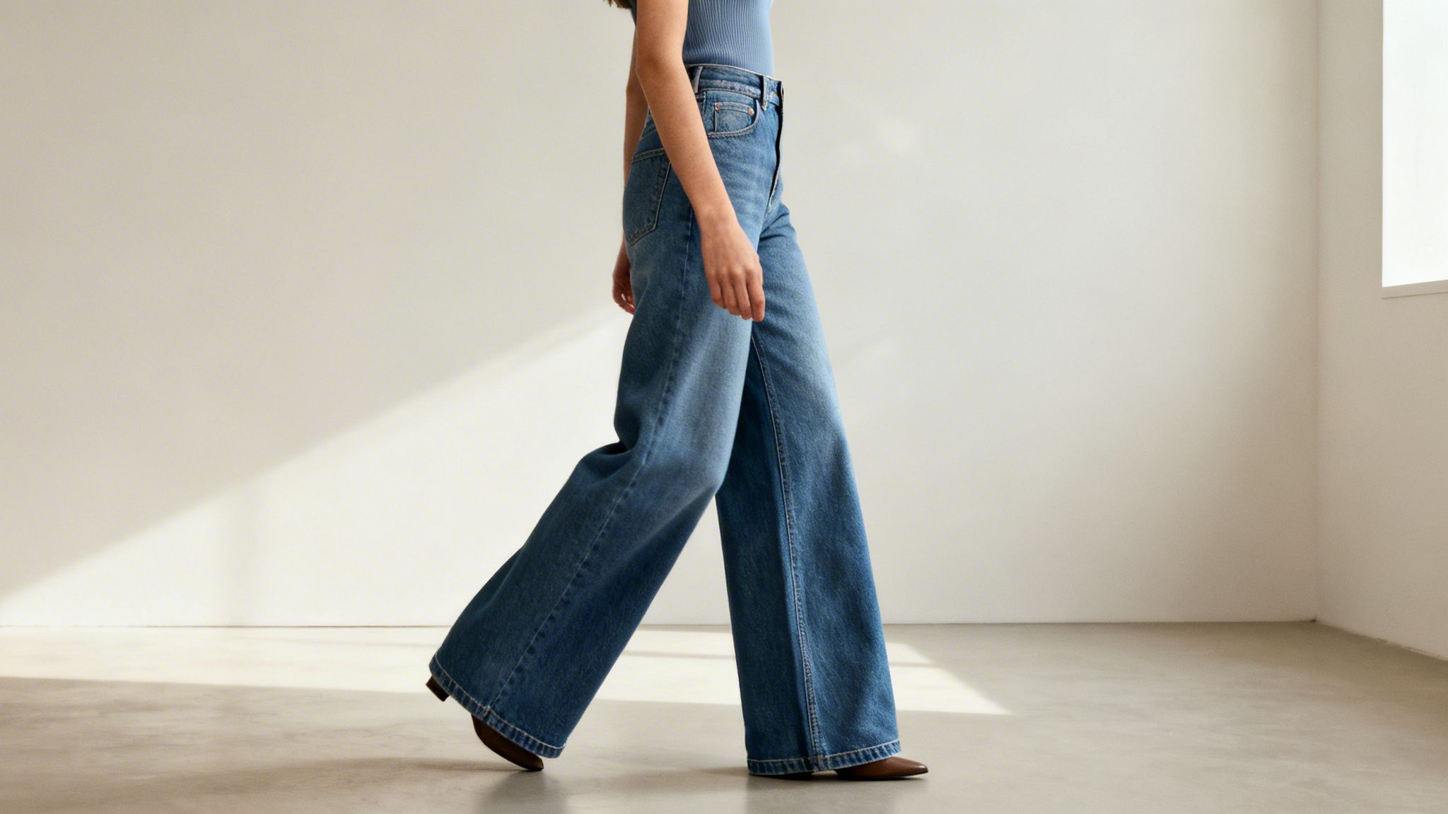 Osoba w niebieskich jeansach z szerokimi nogawkami i brązowych butach idąca w jasnym pomieszczeniu. Światło słoneczne tworzy cień na ścianie.