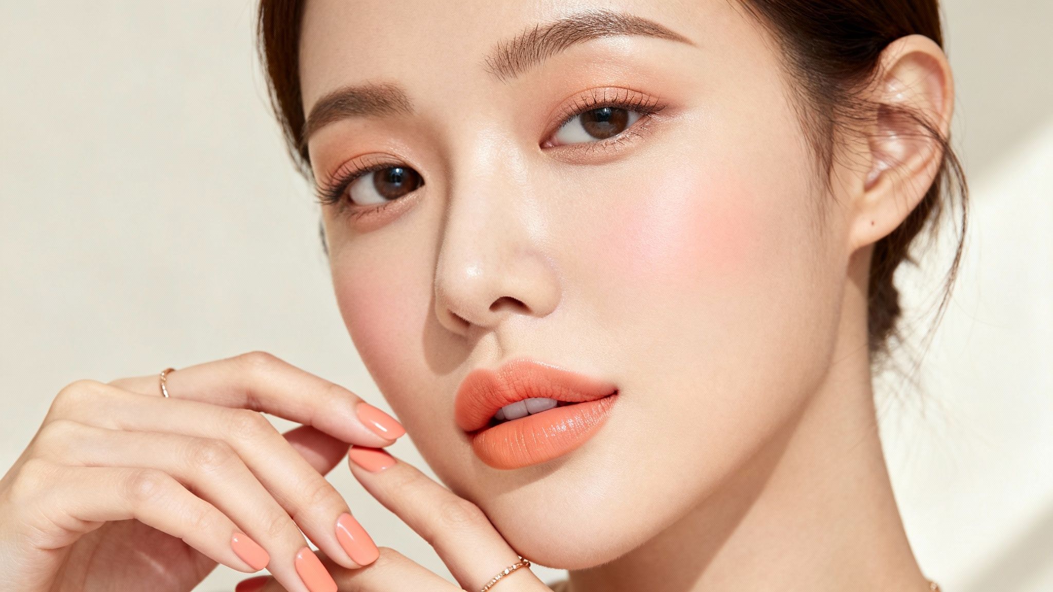 Zbliżenie na twarz i dłonie kobiety z makijażem w brzoskwiniowych odcieniach, z pomarańczową szminką i lakierem do paznokci.