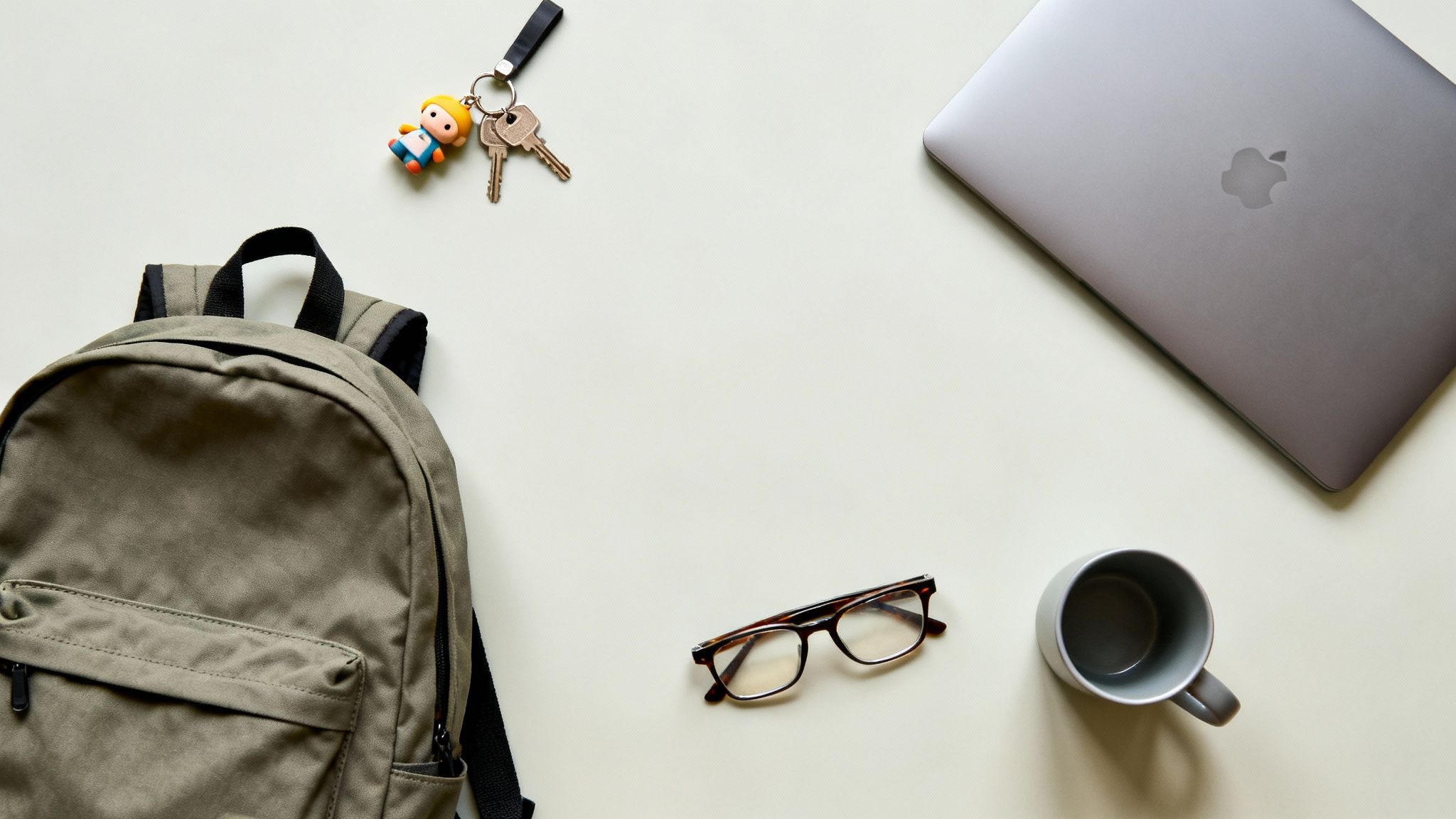 Płaskie ułożenie przedmiotów codziennego użytku: plecak, klucze z breloczkiem, laptop, okulary i kubek na jasnym tle.