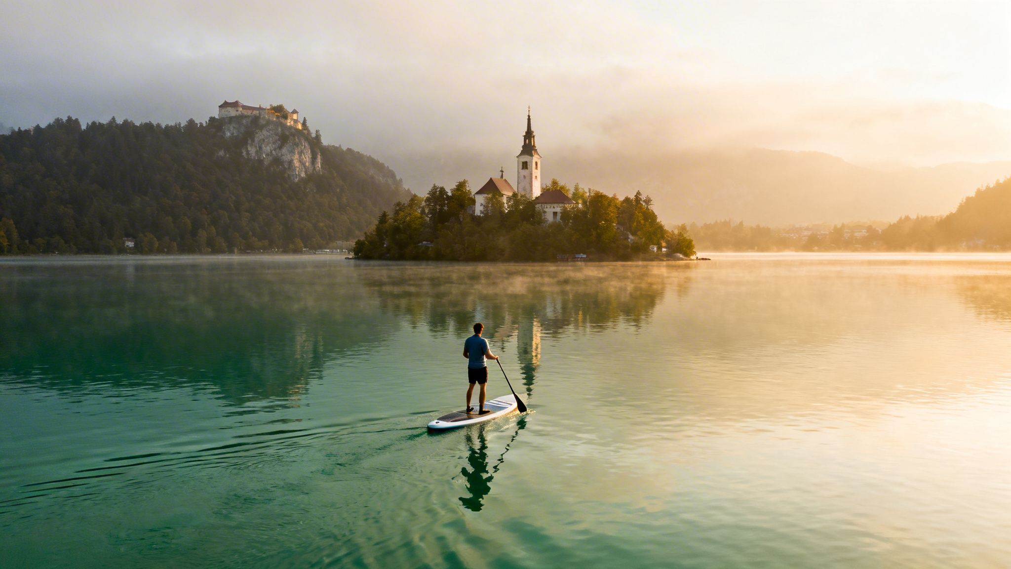 A person paddleboarding on a serene, misty lake towards a church on an island at sunrise.