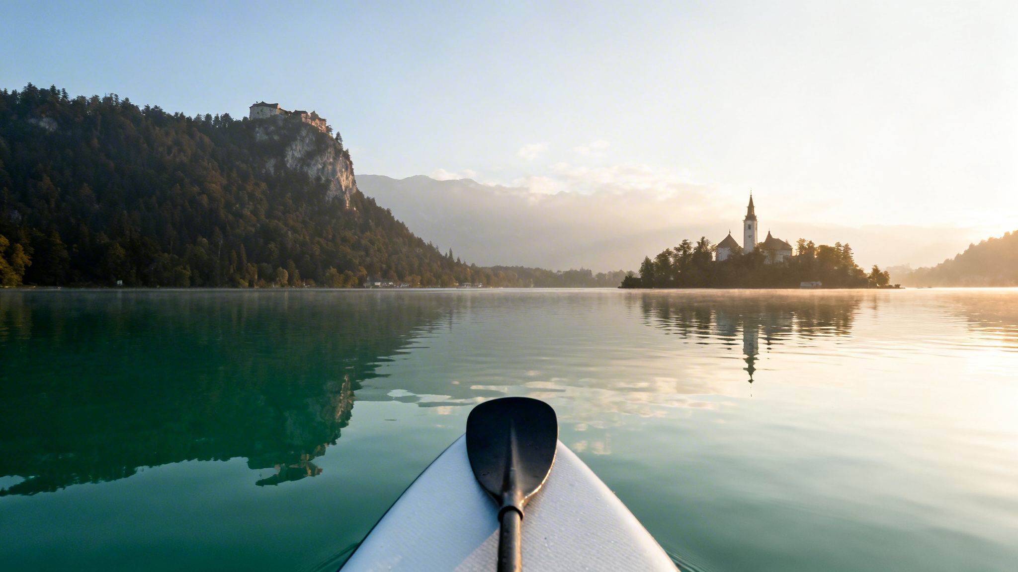 Paddleboard perspective of Lake Bled at sunrise, with the church island and castle in the distance.
