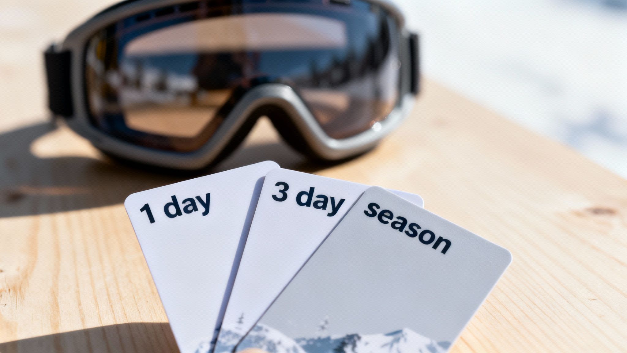 Three white ski passes for 1 day, 3 days, and season next to ski goggles on wood.