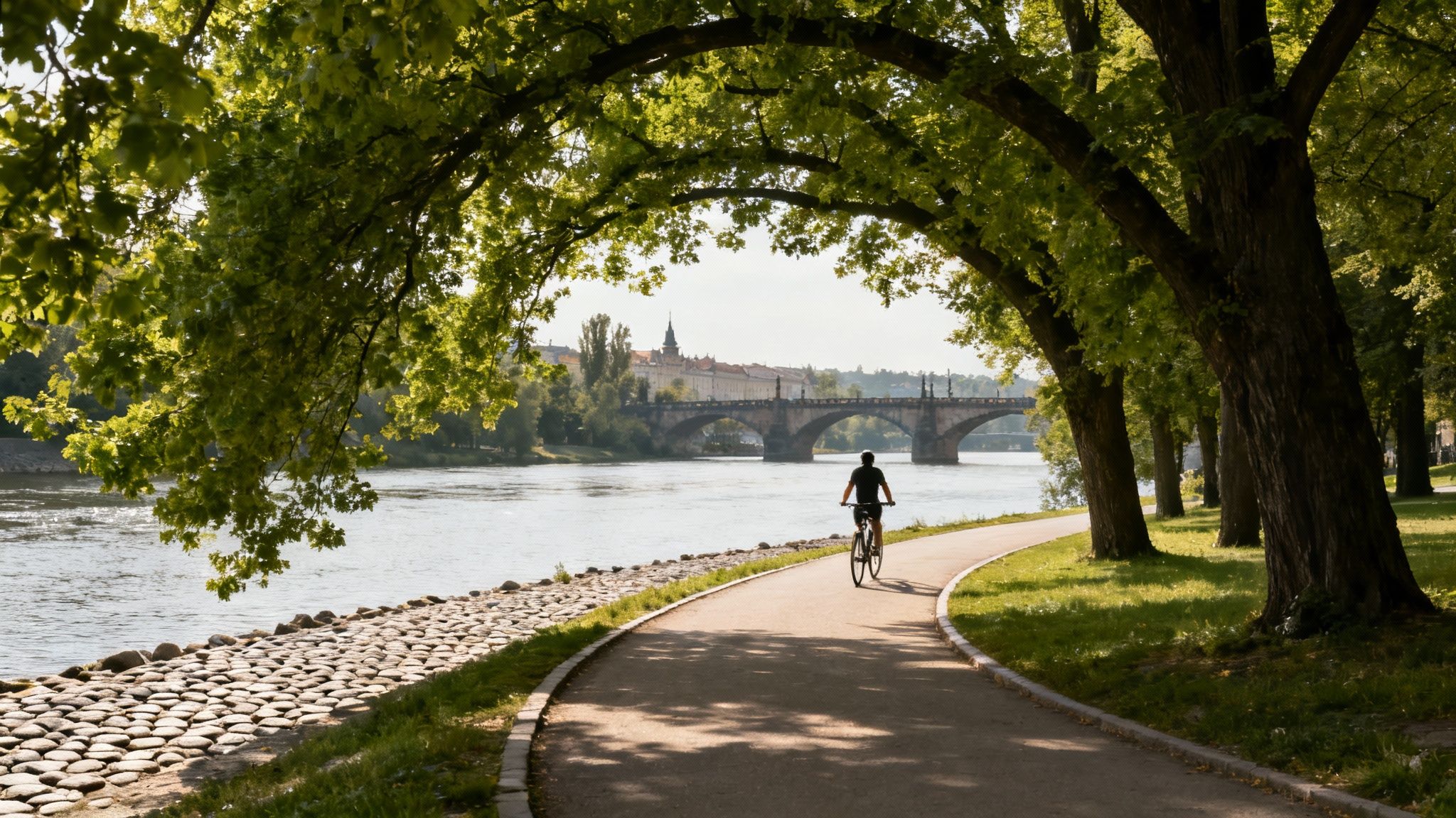 A person cycles on a scenic riverside path under green tree arch, with a historic bridge and city in the background.
