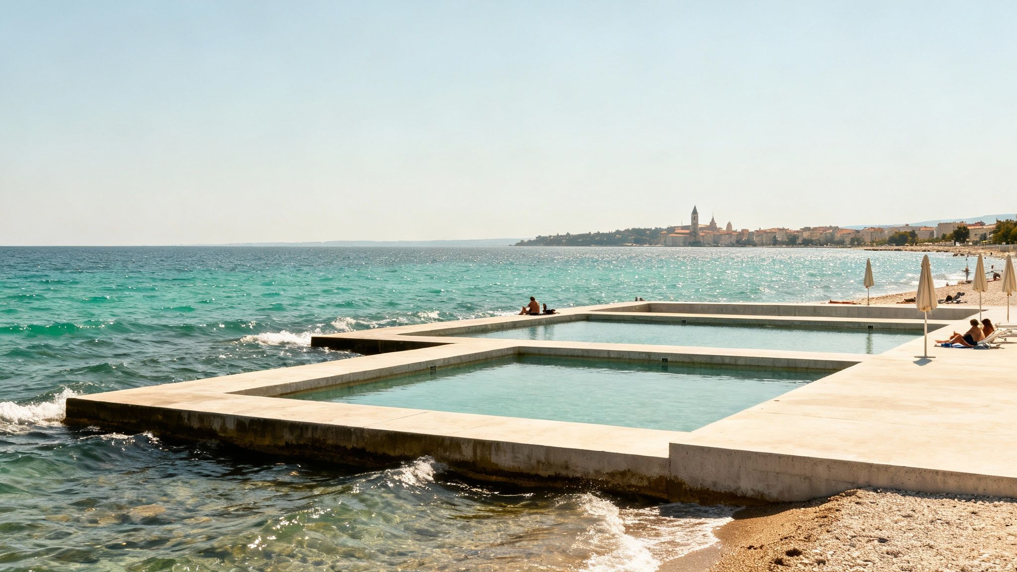 Concrete sea pools on a sunny beach, with people relaxing and a coastal town in the distance.