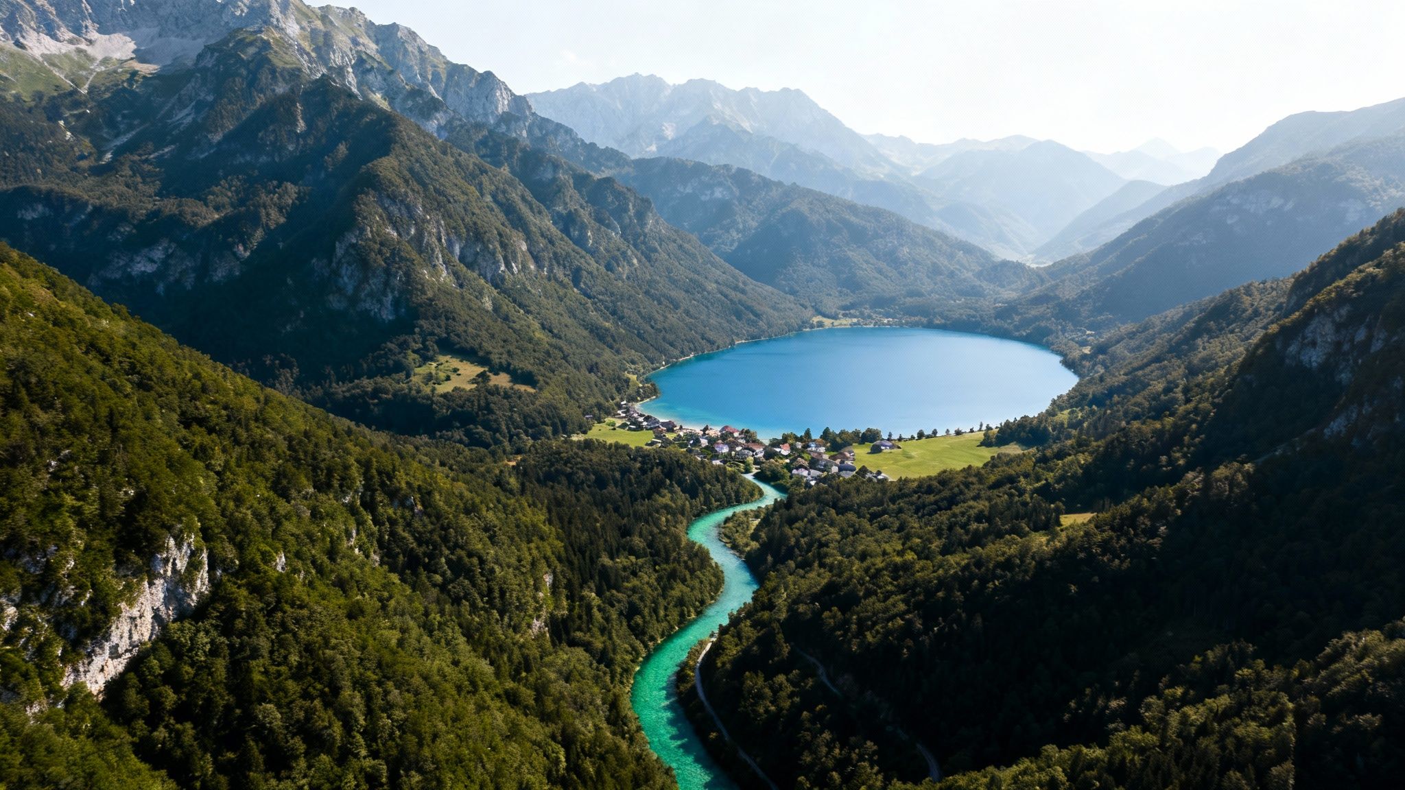 Aerial view of a turquoise river leading to a blue lake, village, and green mountains.
