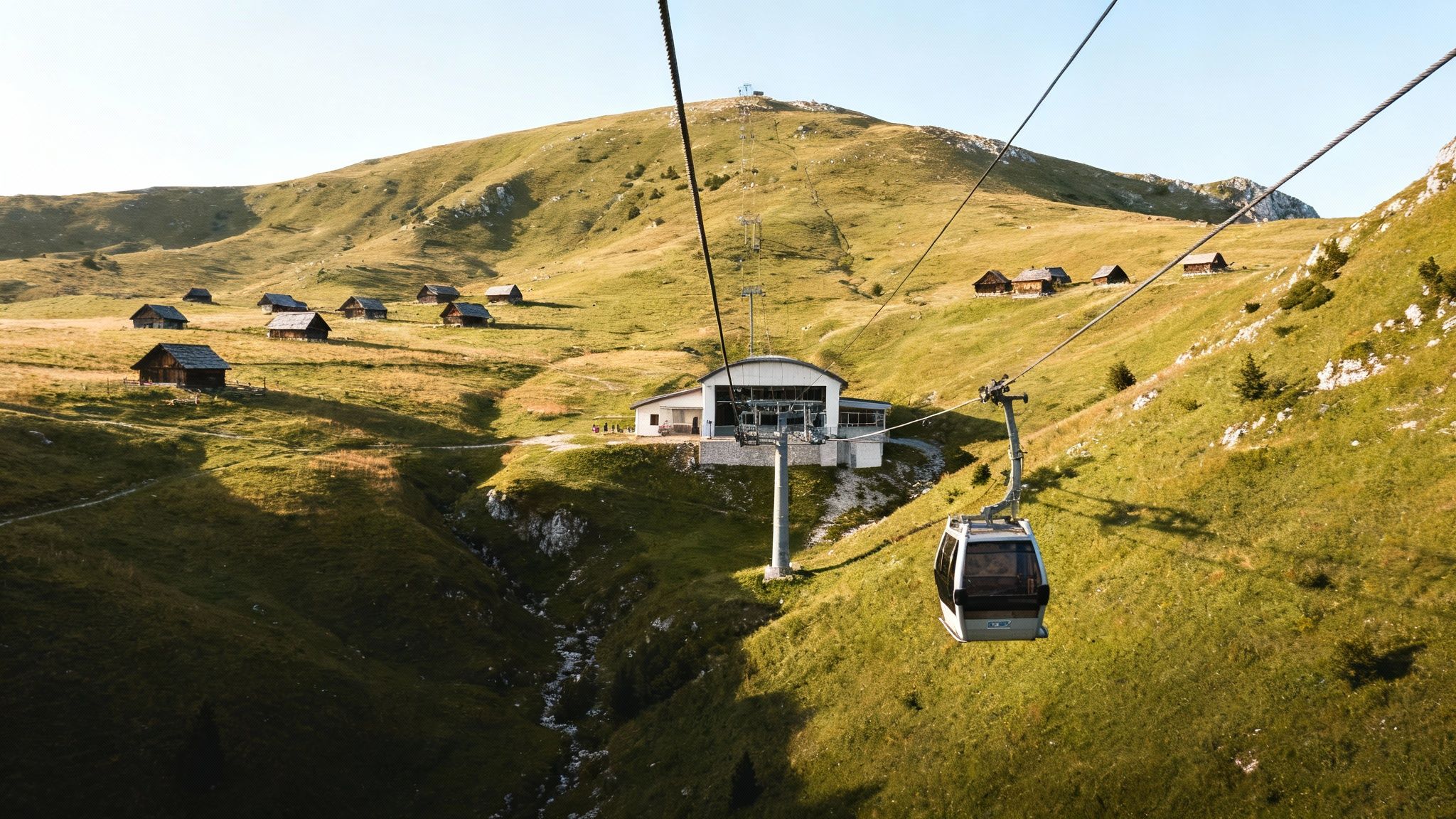 A scenic view of a cable car cabin ascending over a lush green mountain landscape with traditional wooden huts.