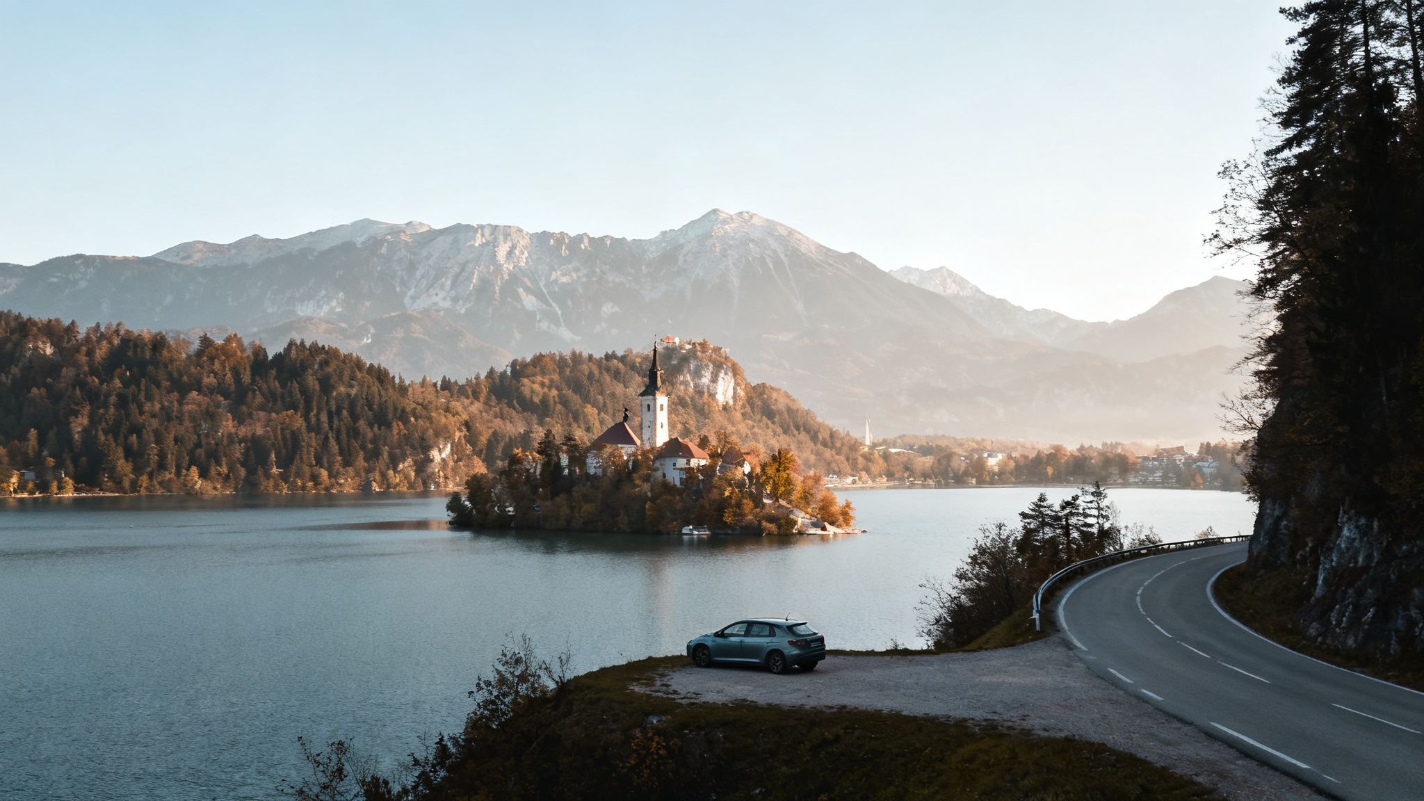 Panoramic view of Lake Bled with its island church, snowy mountains, and a car on a winding road.