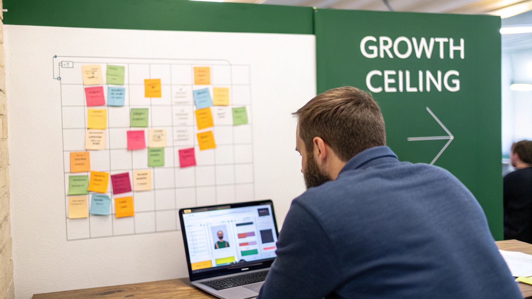 A man works on a laptop in a modern office with a whiteboard full of colorful sticky notes.