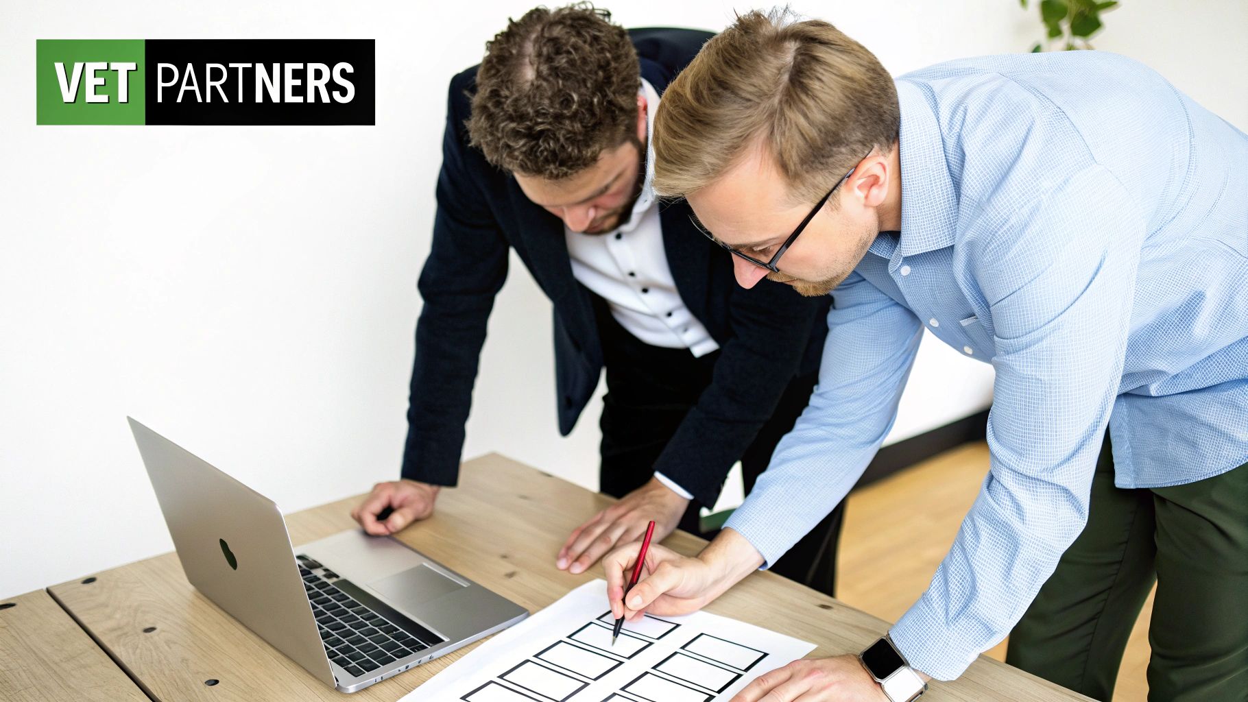Two men collaborating on a document with rectangular frames on a wooden table, with a laptop nearby.