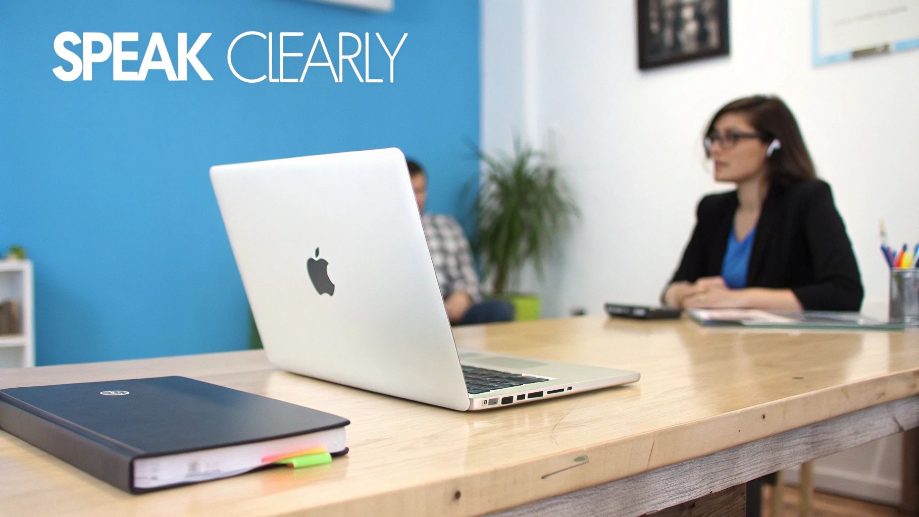 A MacBook on a wooden desk with a notebook, and people in the background, promoting clear speech.