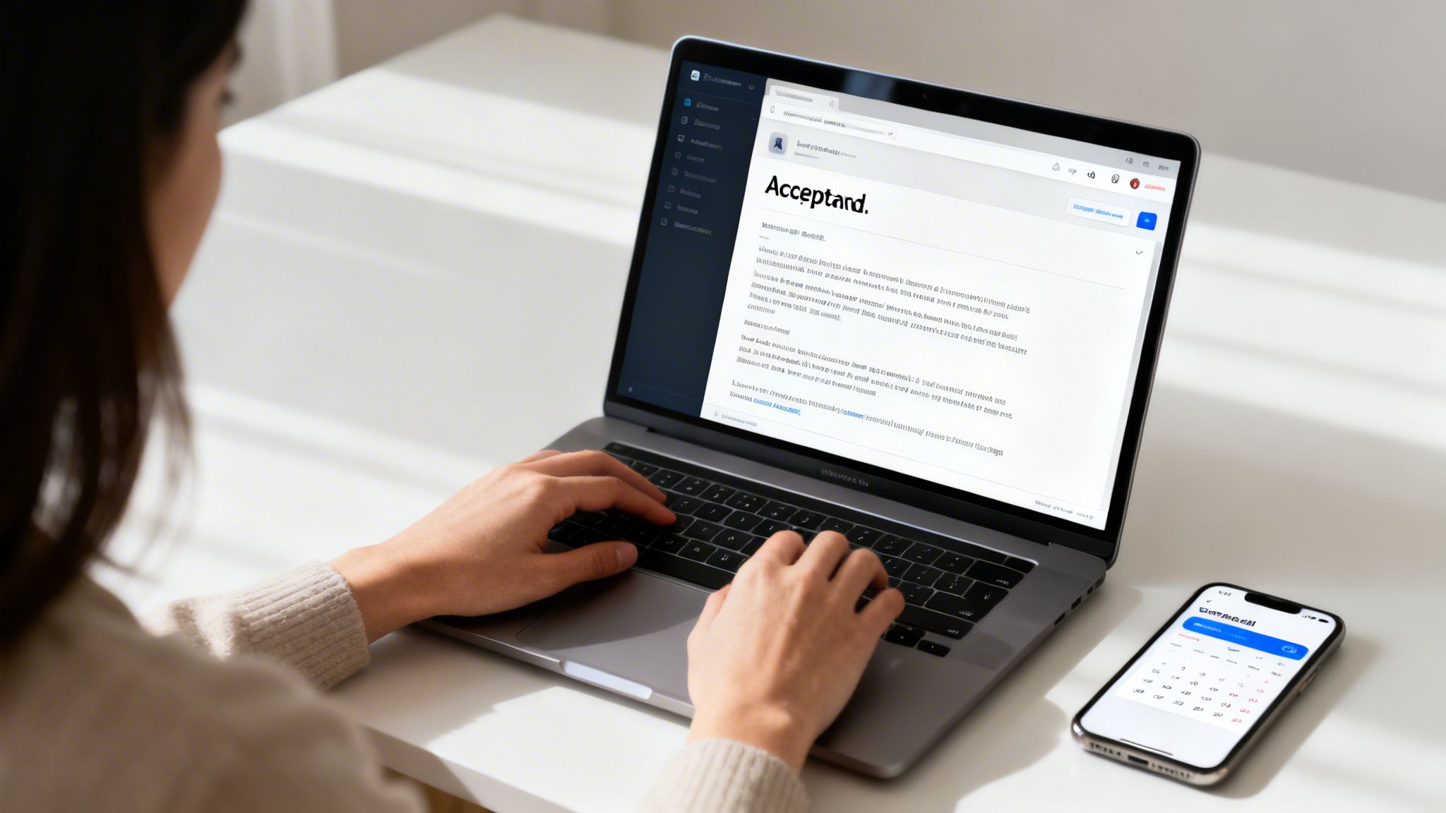 A person typing on a laptop displaying a document titled 'Acceptand.', with a smartphone showing a calendar nearby.