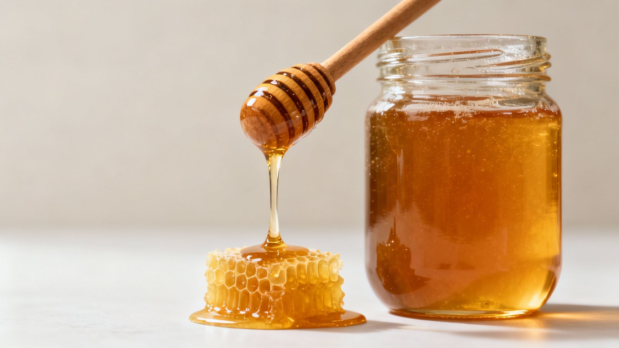 A wooden honey dipper drips golden honey onto a honeycomb next to a full honey jar.