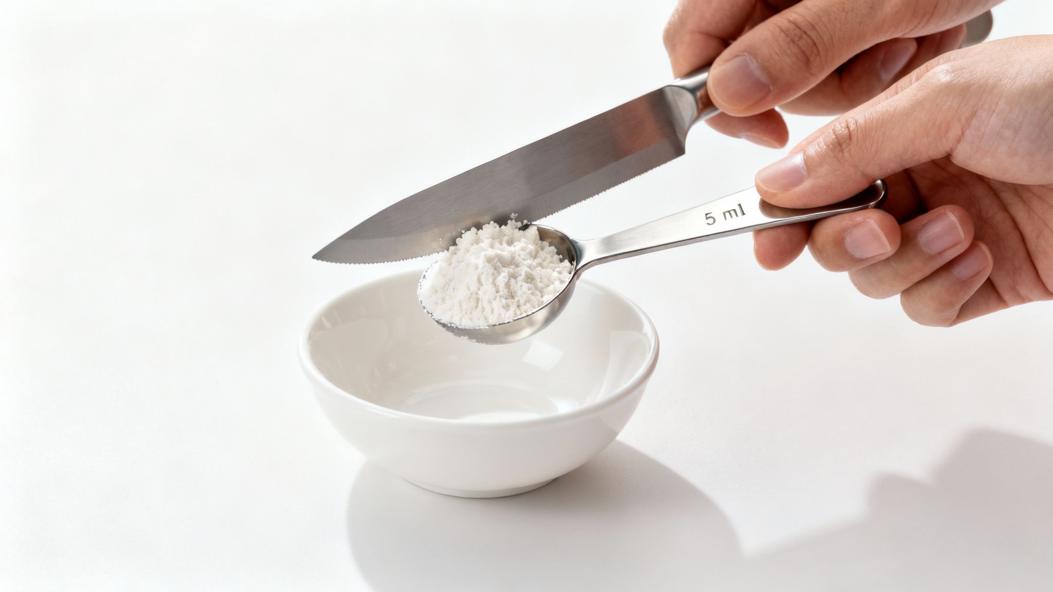 Hands leveling a 5 ml measuring spoon filled with white powder using a knife, above a white bowl.
