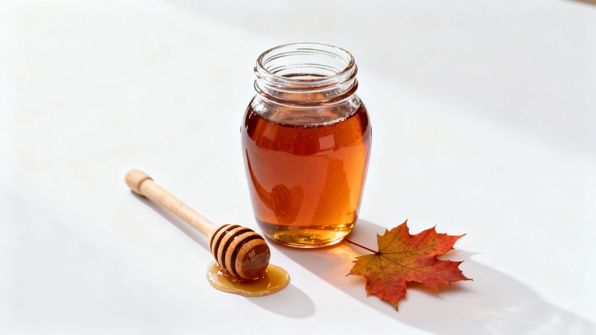 A clear glass jar of golden maple syrup with a wooden dipper and a colourful autumn leaf.