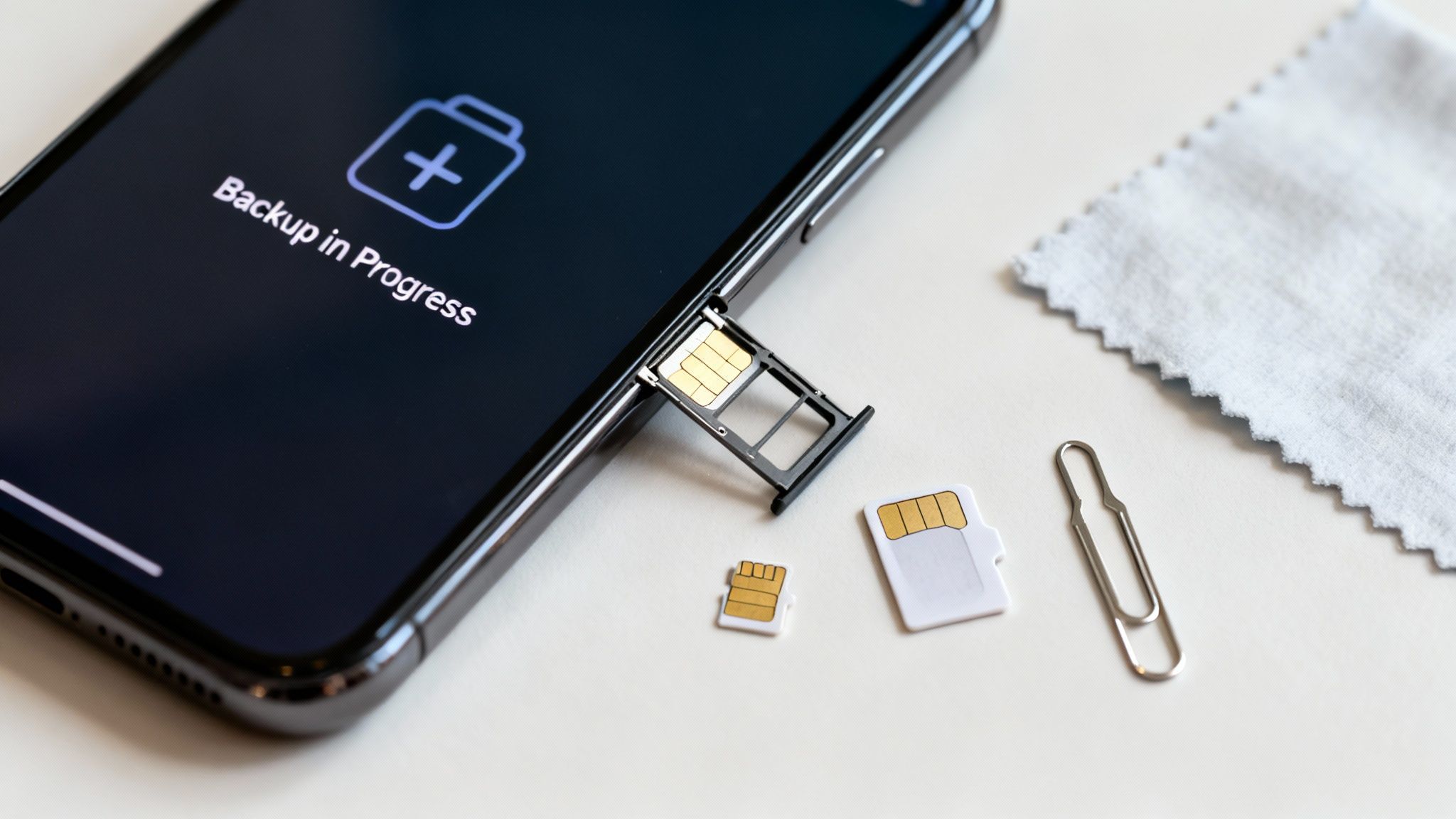 A smartphone displays 'Backup in Progress' with an ejected SIM tray, SIM cards, and a paperclip nearby.