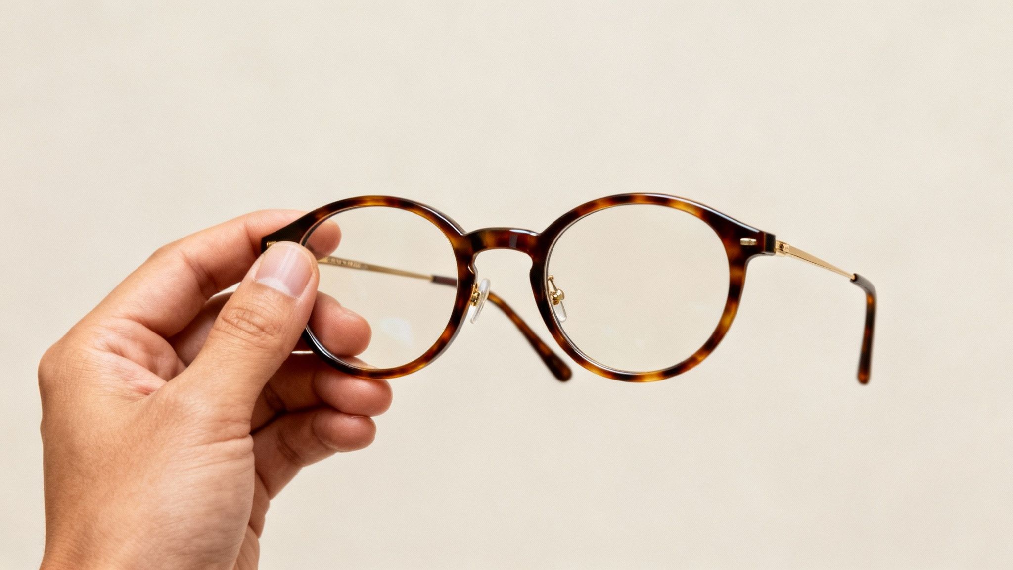 Hand holding tortoiseshell round eyeglasses with gold temples against neutral beige background