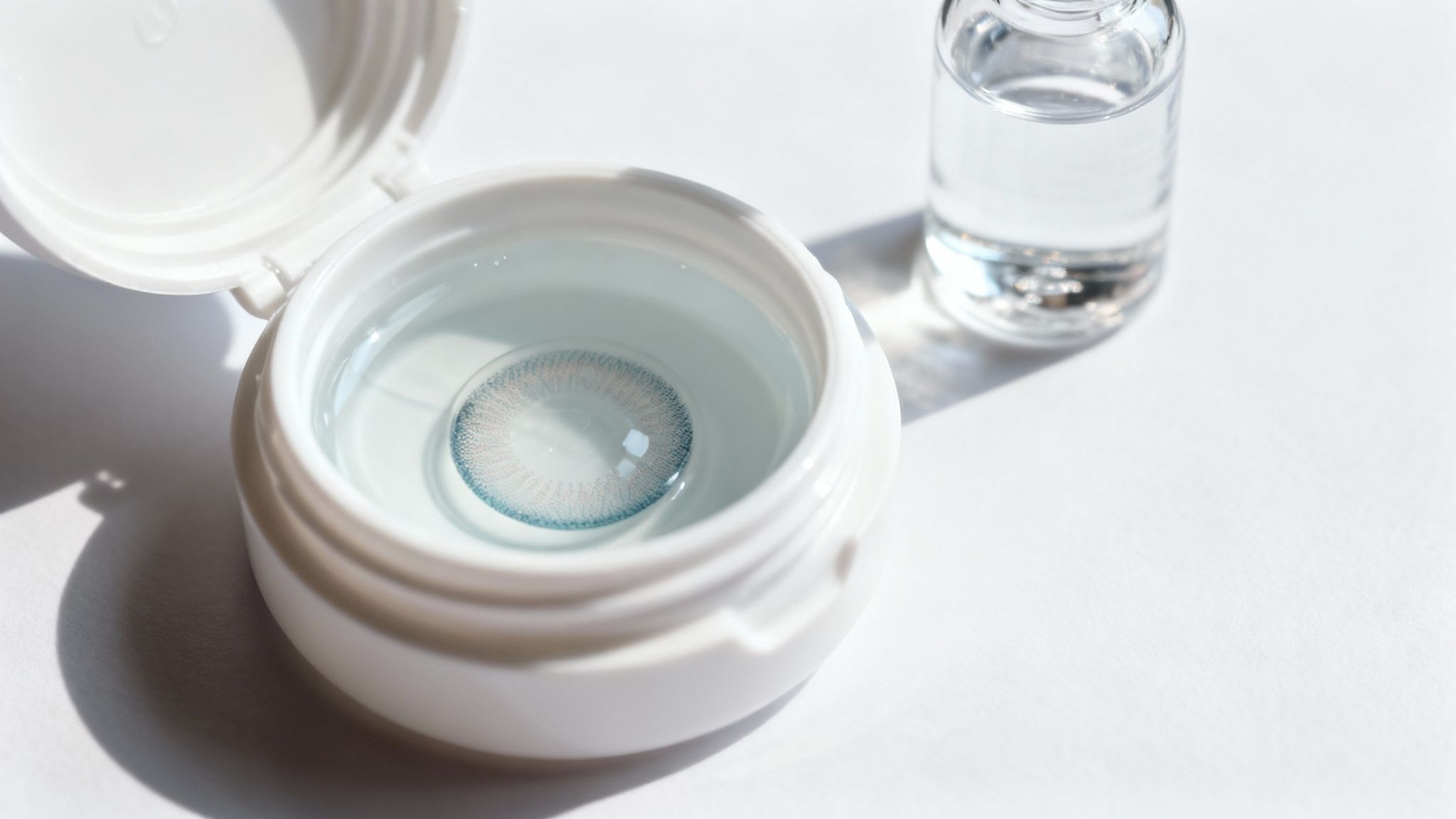 Blue contact lens soaking in clear solution inside white storage case with water bottle