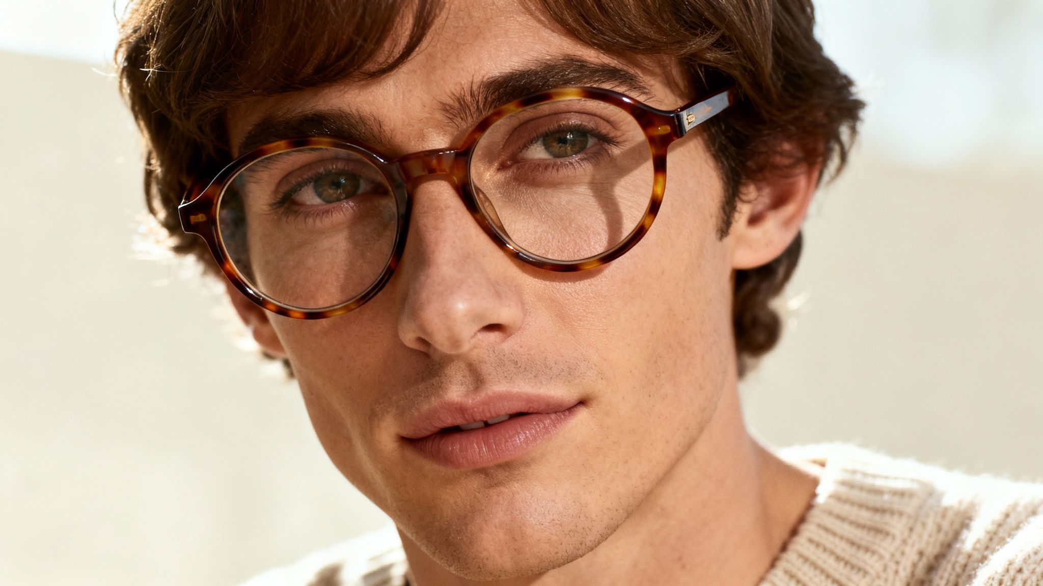 Close-up of a man with brown hair wearing stylish tortoiseshell round eyeglasses and a cream sweater.