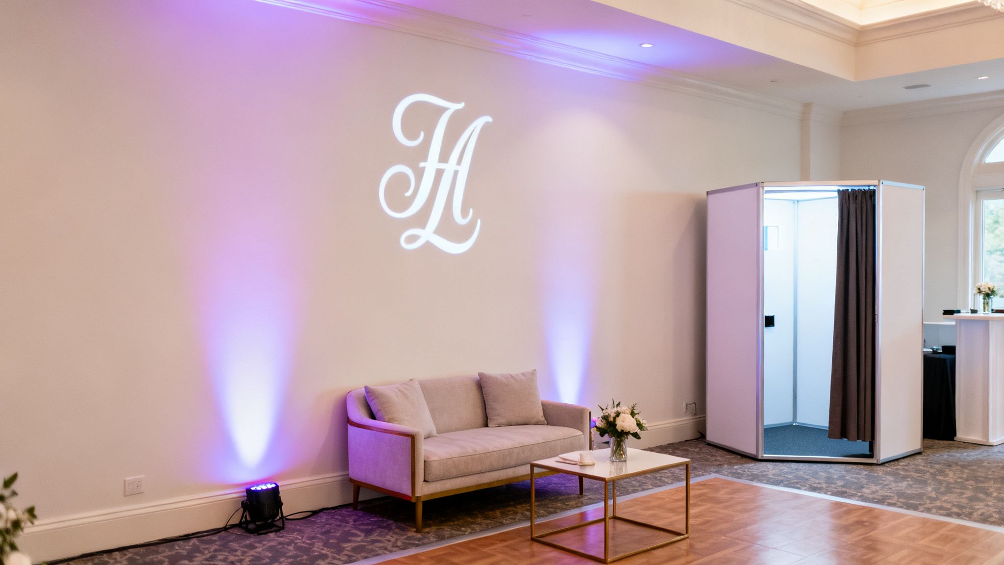 An elegant wedding reception setup with a white photo booth, a beige lounge sofa, and custom monogram lighting.