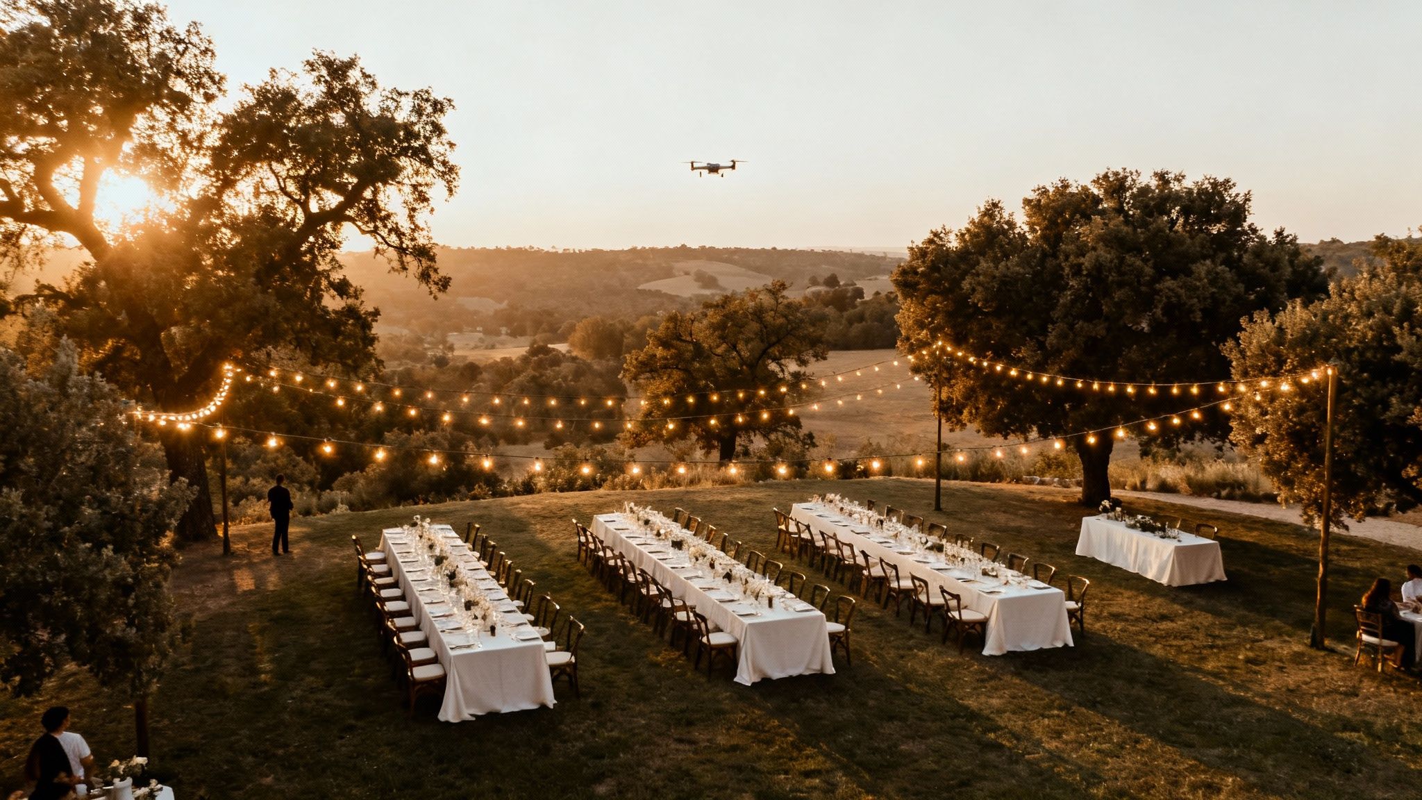 An elegant outdoor dinner party at sunset, with long tables, string lights, and a drone flying.