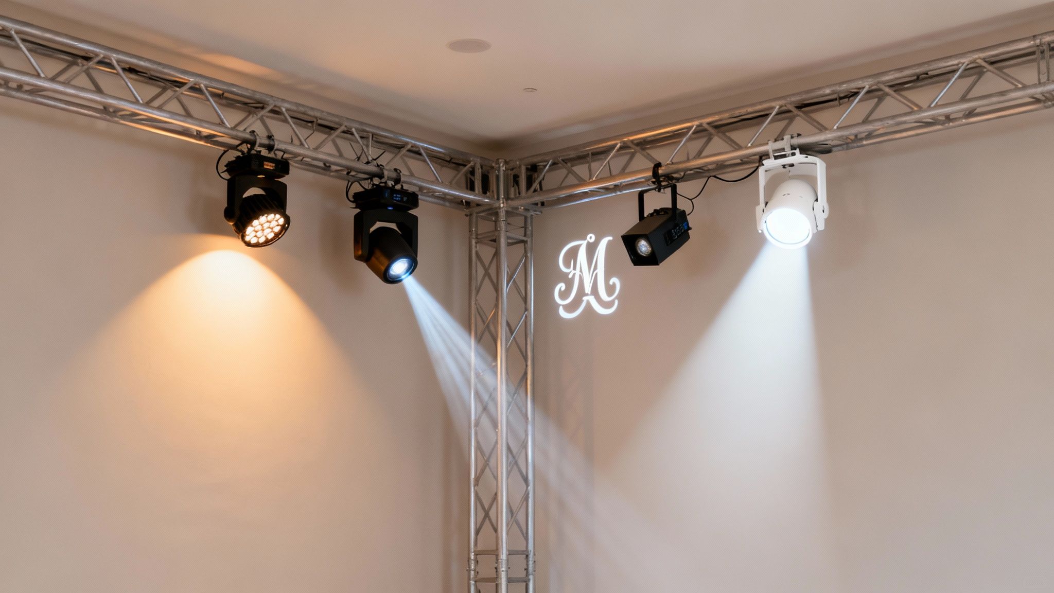 Modern stage lights mounted on a truss system projecting light and an 'M' logo onto a wall.