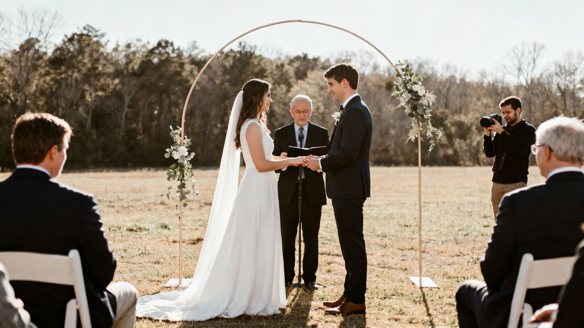 An outdoor wedding ceremony with a bride and groom holding hands under a golden arch, surrounded by guests.