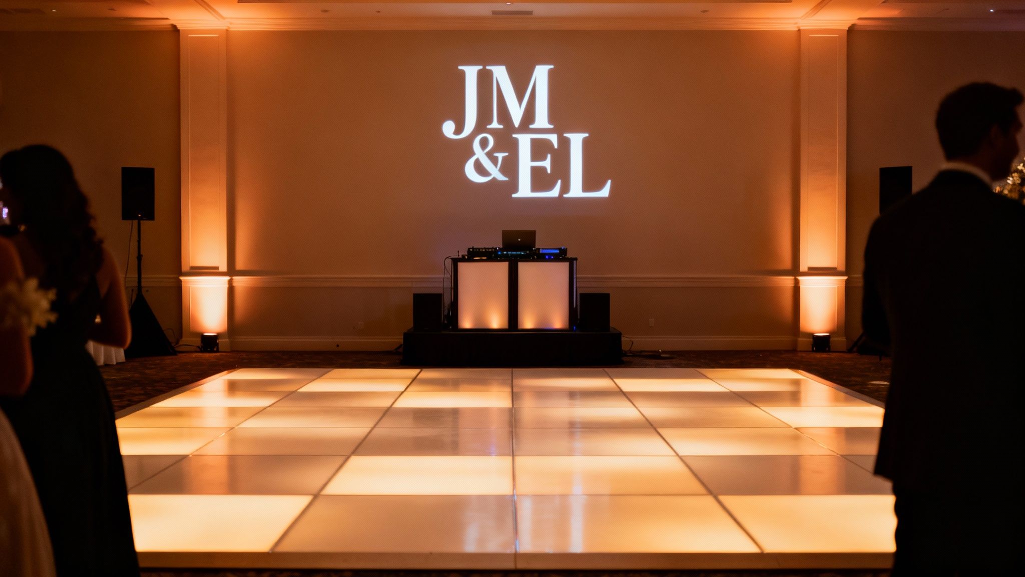 Wedding reception hall with illuminated dance floor, DJ setup, and initials projected on the wall.