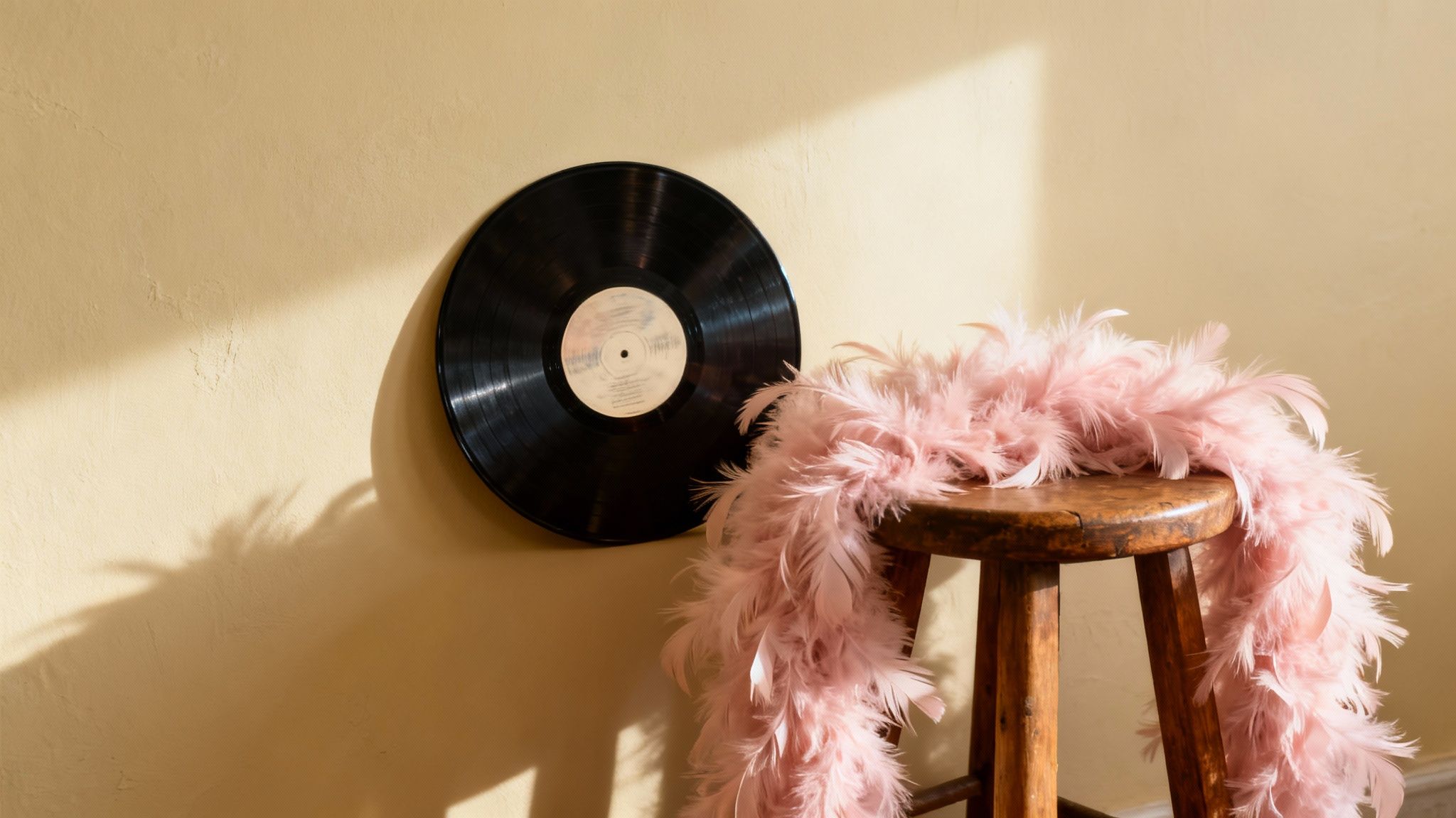 A pink feather boa draped on a rustic wooden stool next to a vinyl record on a yellow wall, bathed in sunlight.