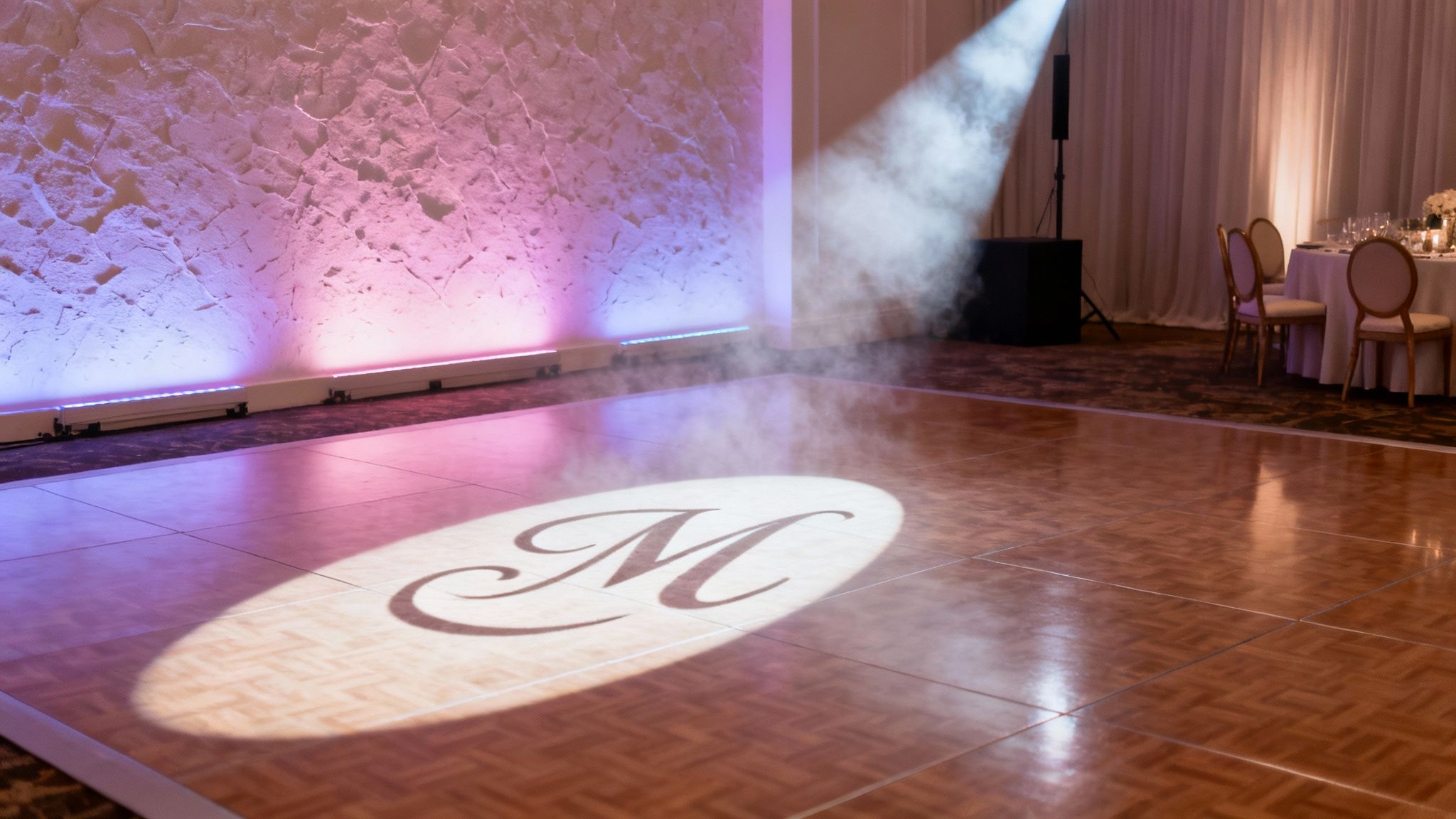 A wedding reception dance floor with a monogram 'M' projected, uplighting, and atmospheric fog.