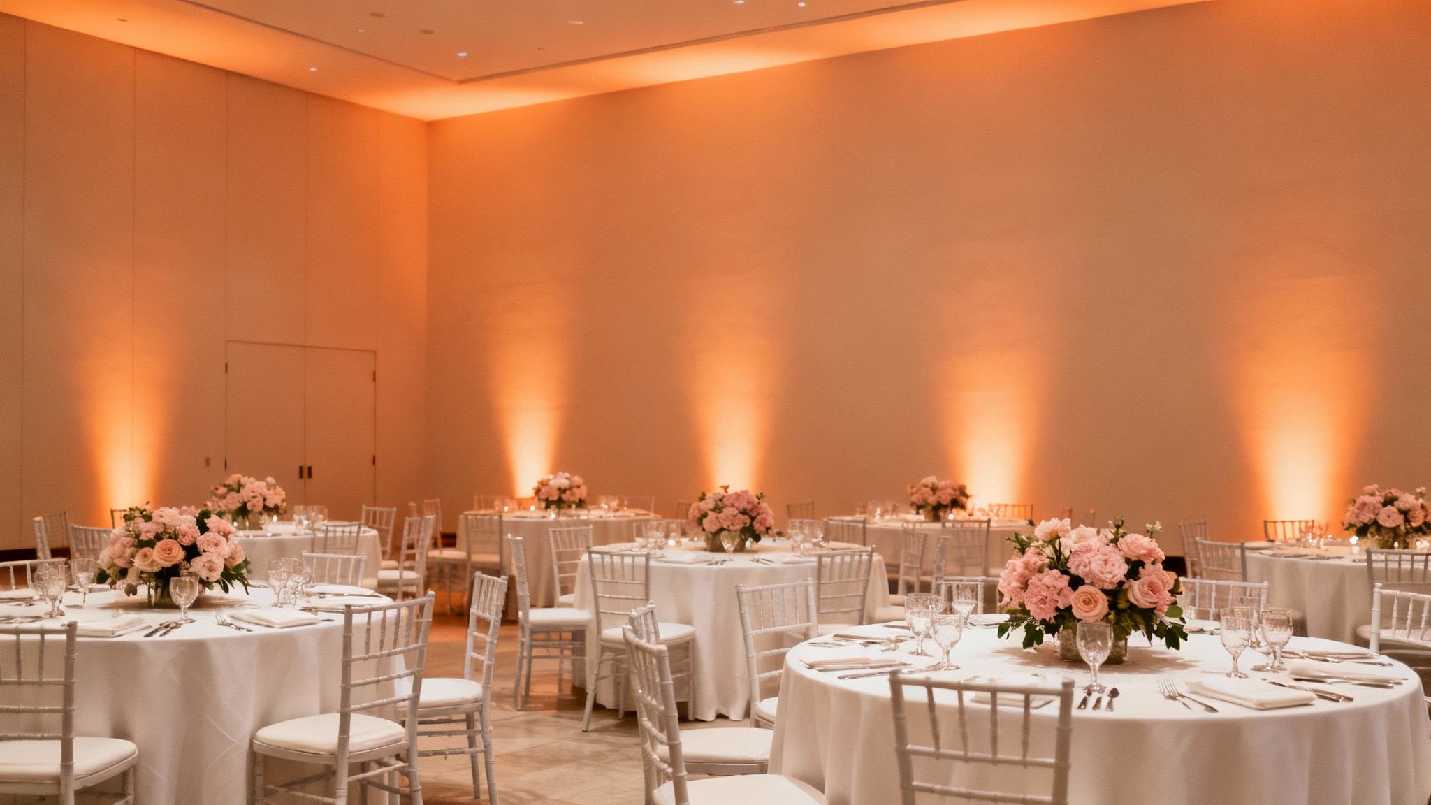 A banquet room with round tables, white chairs, pink floral centerpieces, and warm orange uplighting on the walls.