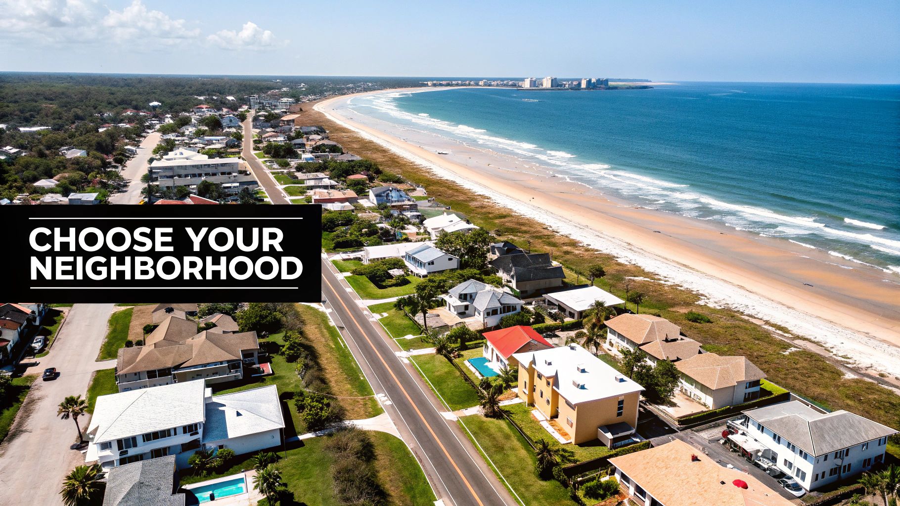 Aerial view of a sunny beachside neighborhood with houses, a road, sandy beach, and blue ocean.