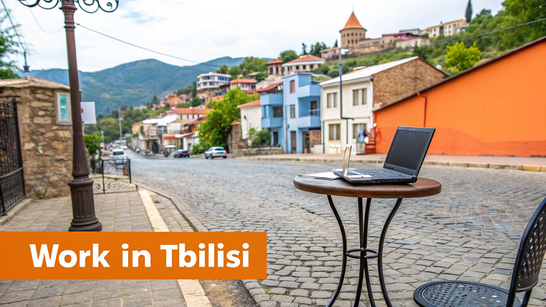 A laptop and notebook on a wooden table on a cobblestone street in Tbilisi, Georgia, with colorful houses and mountains in the background.