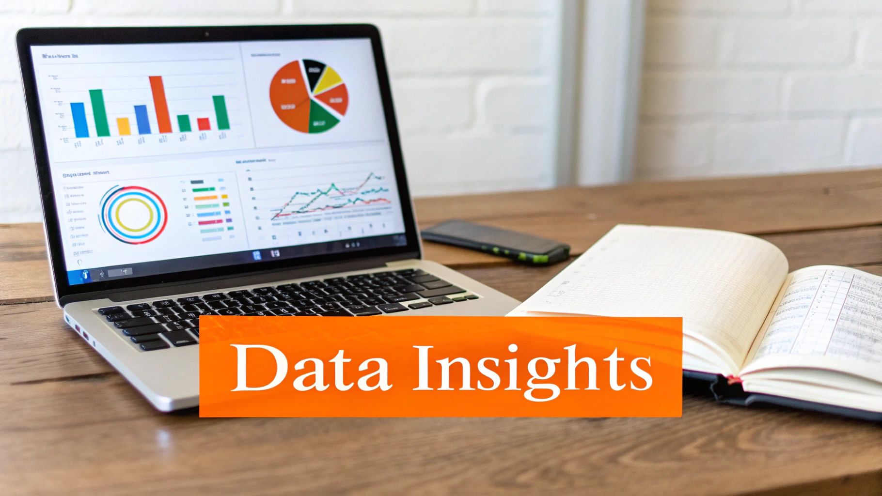 A laptop displaying data insights dashboards with various charts and graphs, a phone, and a notebook on a wooden desk.