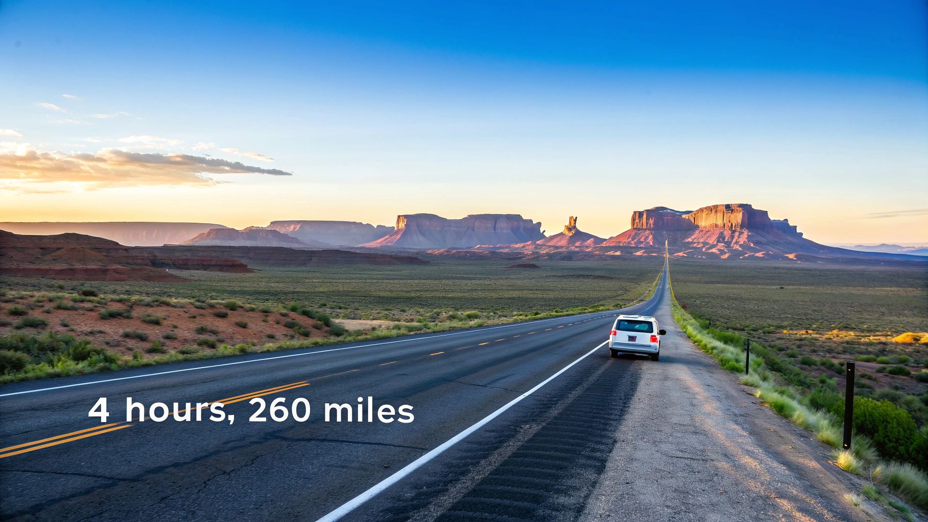 A lone car travels on a long desert road towards distant red rock mountains at sunset.