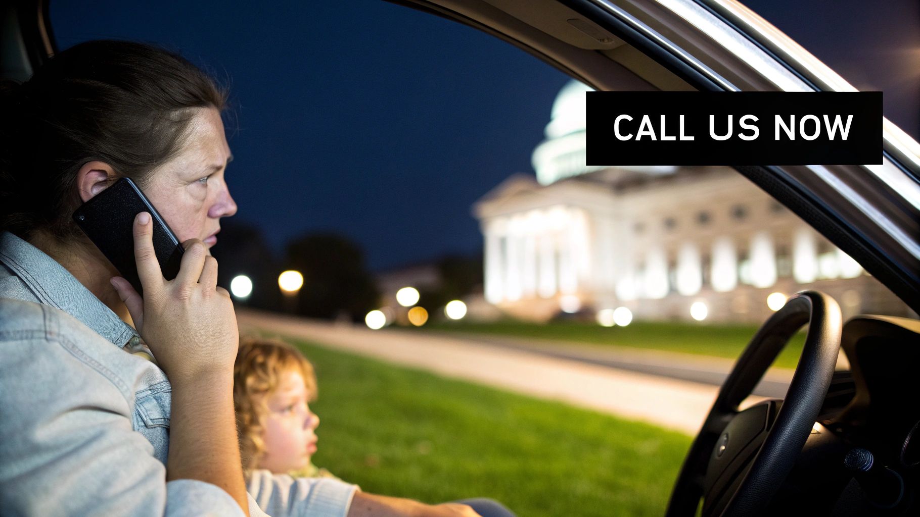A concerned woman on a phone call in a car at night with a child, near a lit government building.